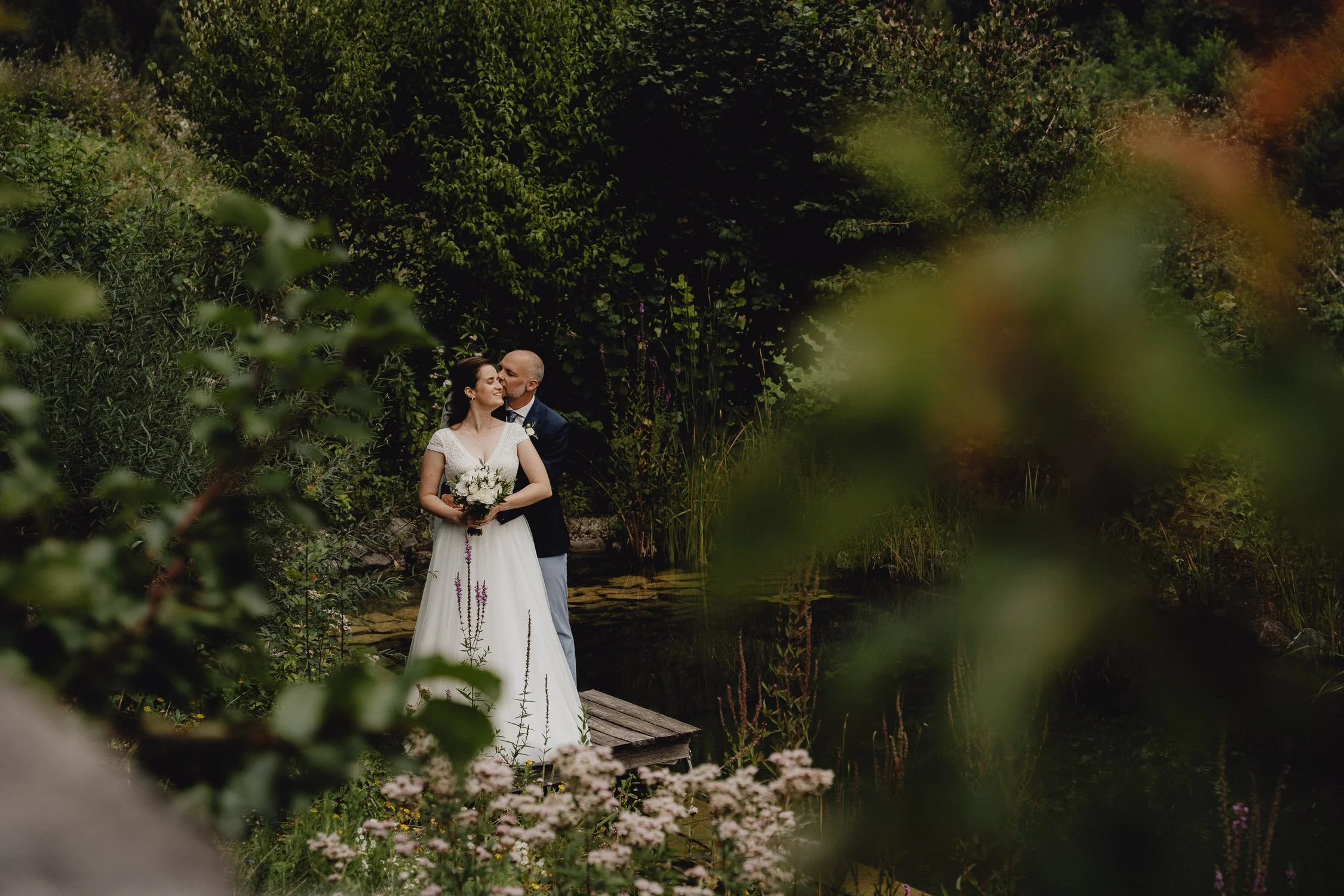 Ein Brautpaar steht auf einem kleinen Holzsteg inmitten eines grünen Gartens, umgeben von Pflanzen und Bäumen, wobei die Frau ein weißes Brautkleid trägt und die Figur des Mannes im Anzug zu sehen ist.