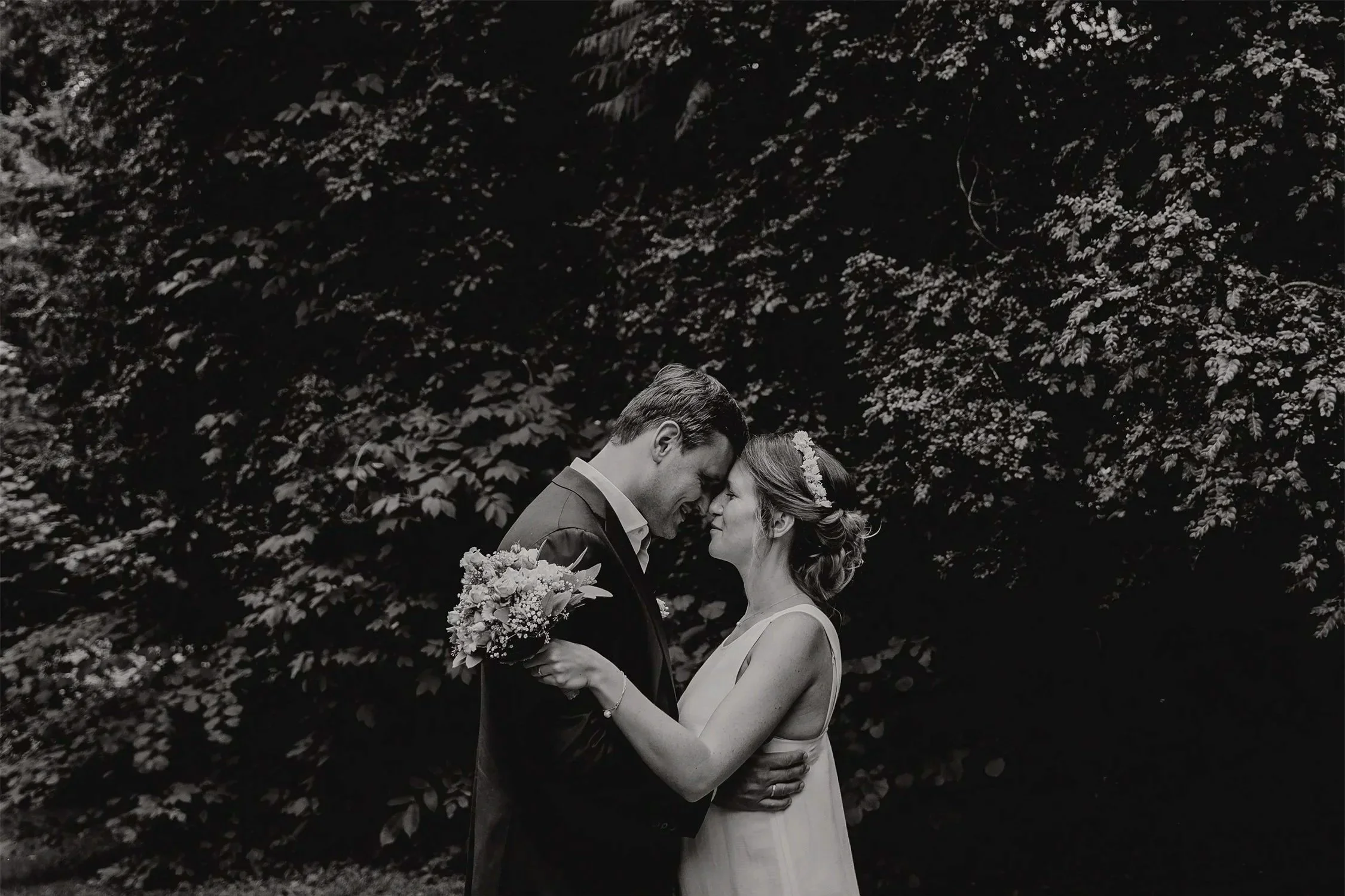 Ein Brautpaar steht sich im Park mit Bäumen im Hintergrund gegenüber, das Brautpaar küsst sich zärtlich, die Braut trägt ein Kleid und eine Haarblume, der Bräutigam einen Anzug, in Schwarz-Weiß. Die Frau hält einen Blumenstrauß.