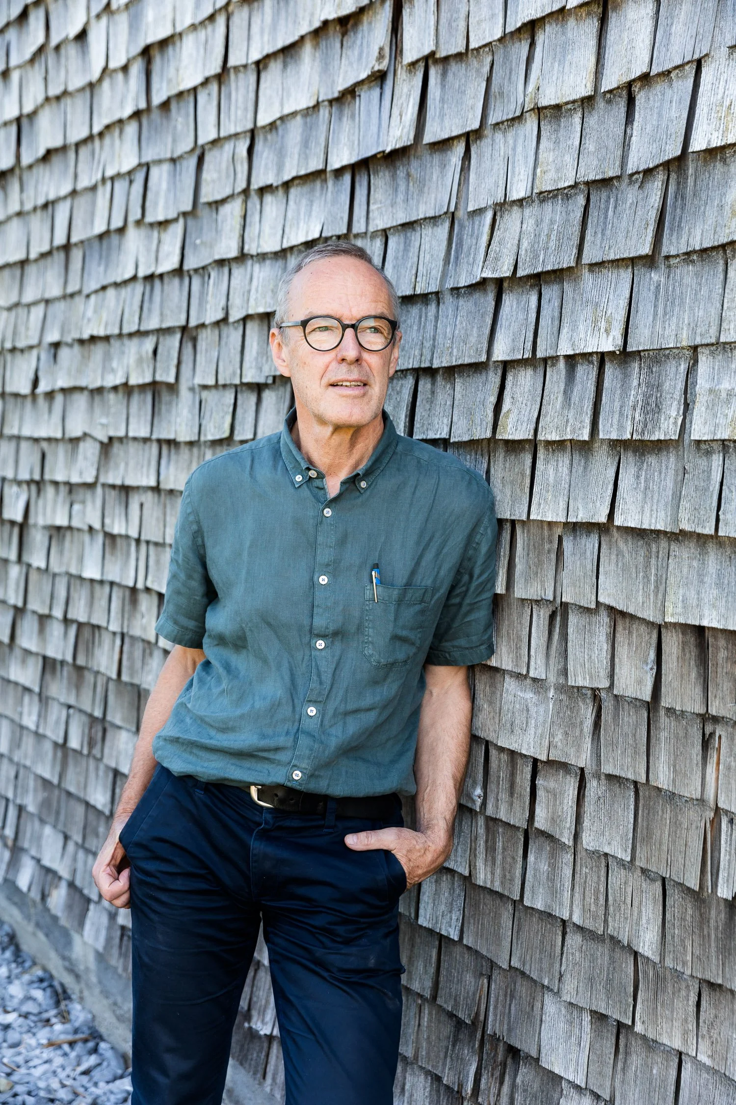 Ein älterer Mann steht vor einer Wand aus Holzschindeln, trägt eine Brille, ein blaugrünes Hemd mit kurzen Ärmeln, schwarze Hose und hat seine rechte Hand in der Hosentasche.