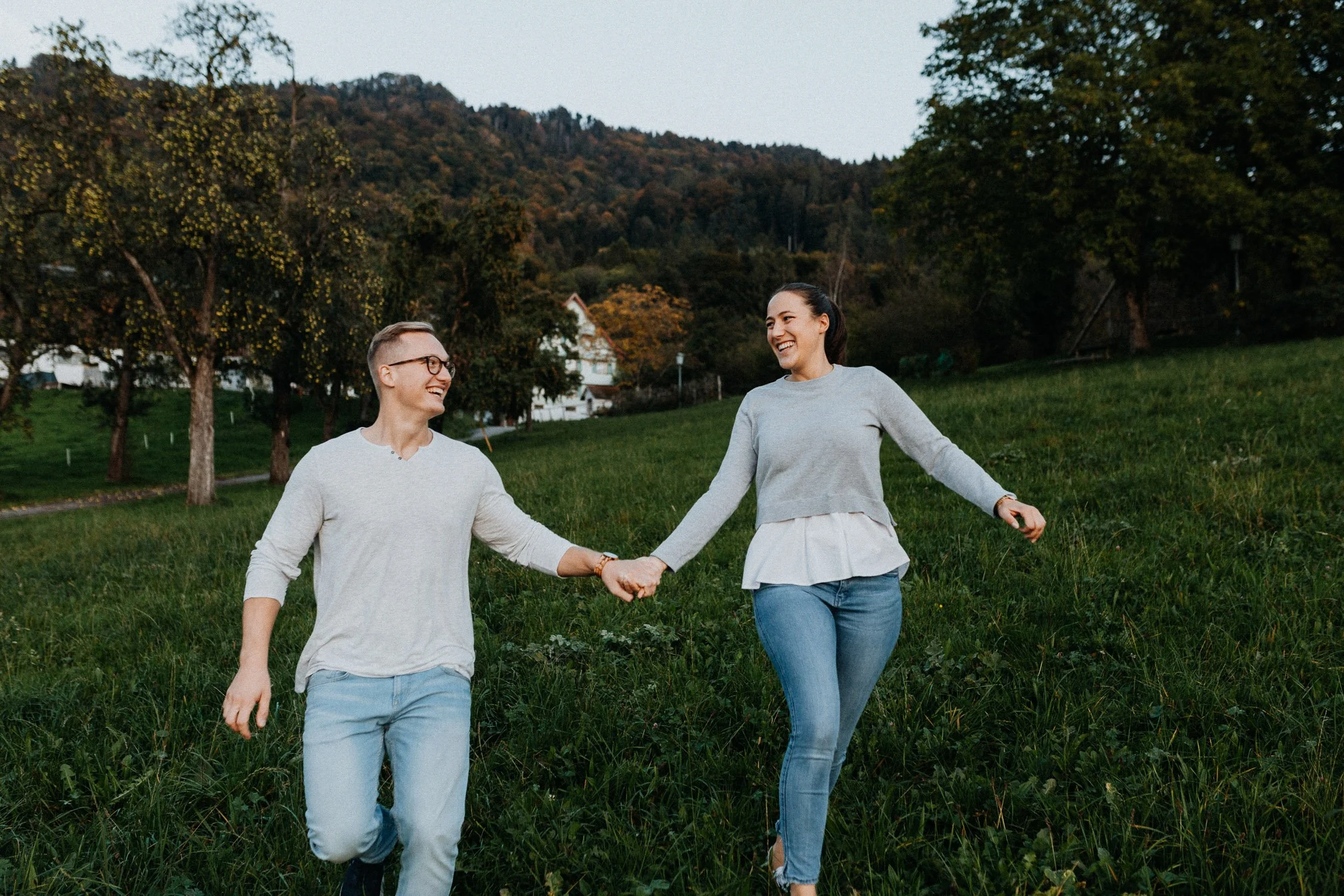 Ein Paar läuft lachend durch eine grüne Wiese, umgeben von Bäumen und Bergen im Hintergrund, bei Sonnenuntergang.