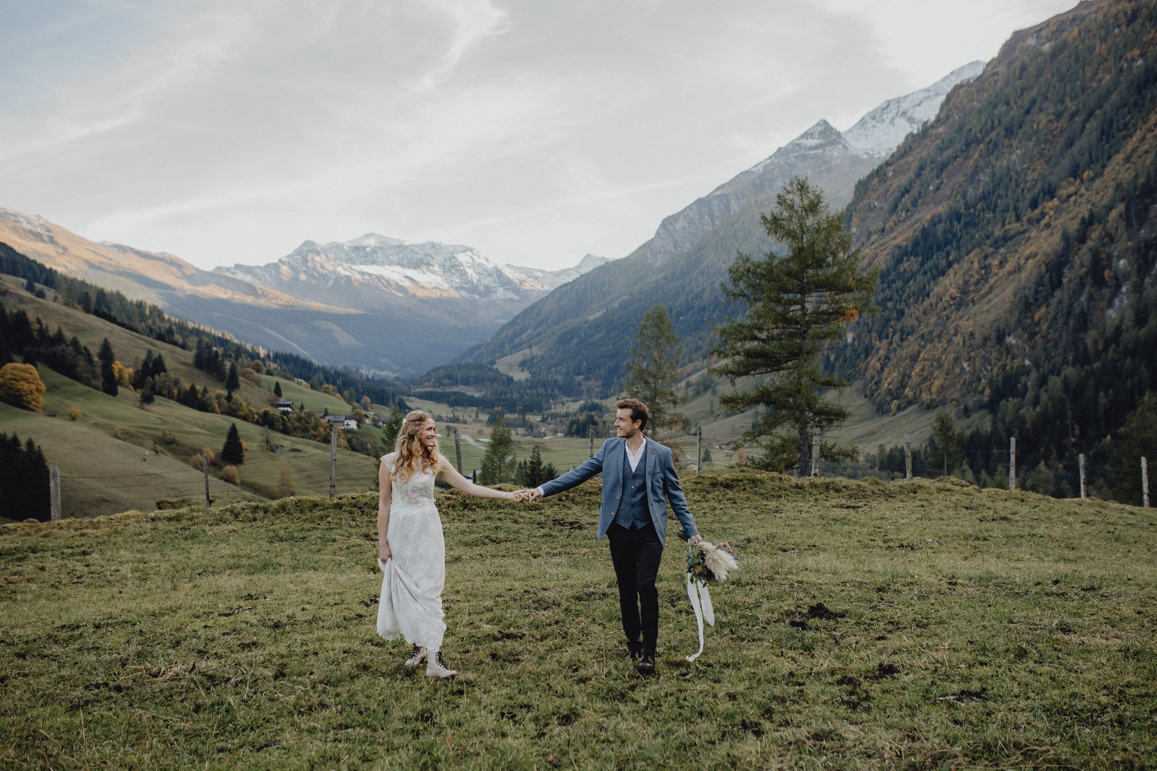 Ein Paar hält sich an einem grünen Berghang in einer malerischen Berglandschaft mit Schneegipfeln im Hintergrund, während sie an einem Tag mit bewölktem Himmel heiraten.