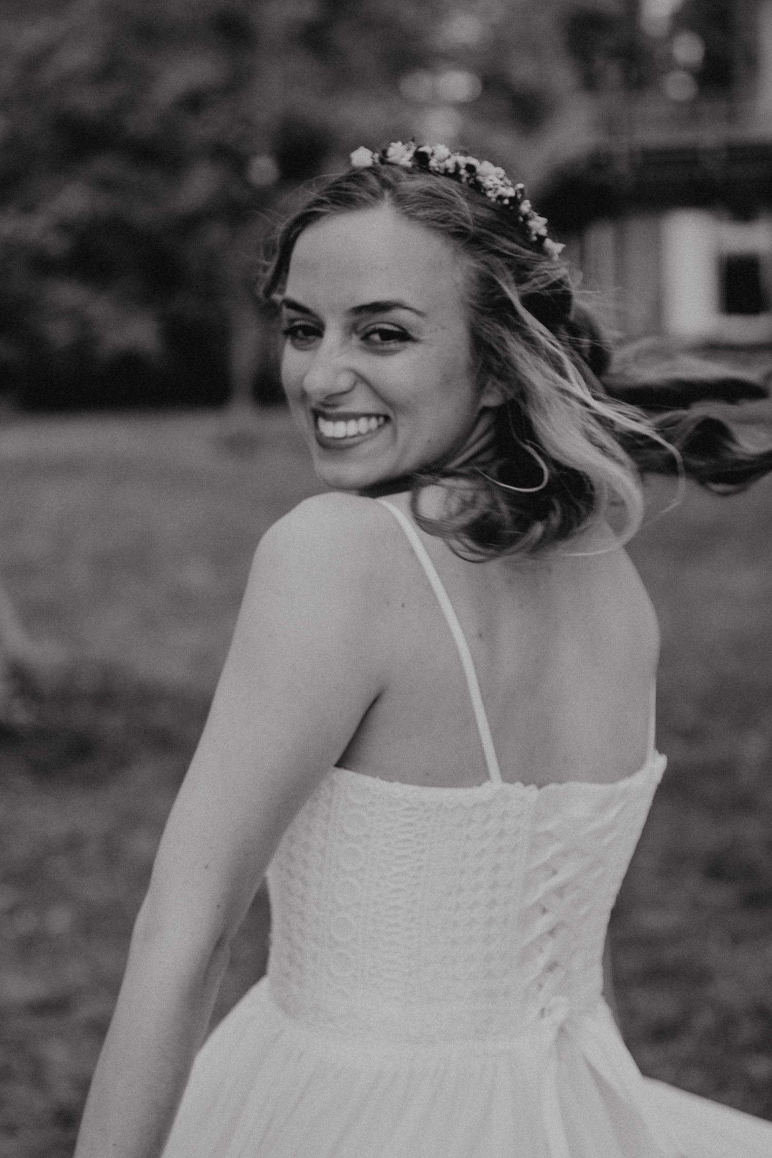 Schwarz-weiß-Foto eines lachenden Mädchens in einem weißen Kleid, das eine Blumenkette trägt, im Freien mit Bäumen und einem Haus im Hintergrund.