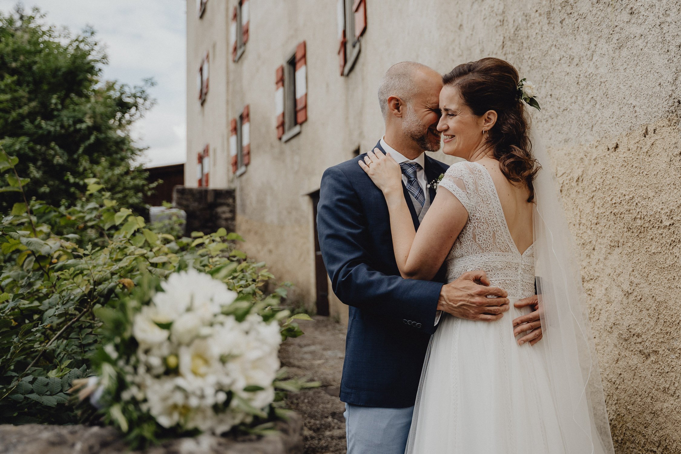 Ein Brautpaar bei einer Hochzeitsaufnahme, umgeben von einem alten Gebäude und Natur. Die Braut trägt ein weißes Kleid mit Spitze, der Bräutigam trägt einen dunklen Anzug.