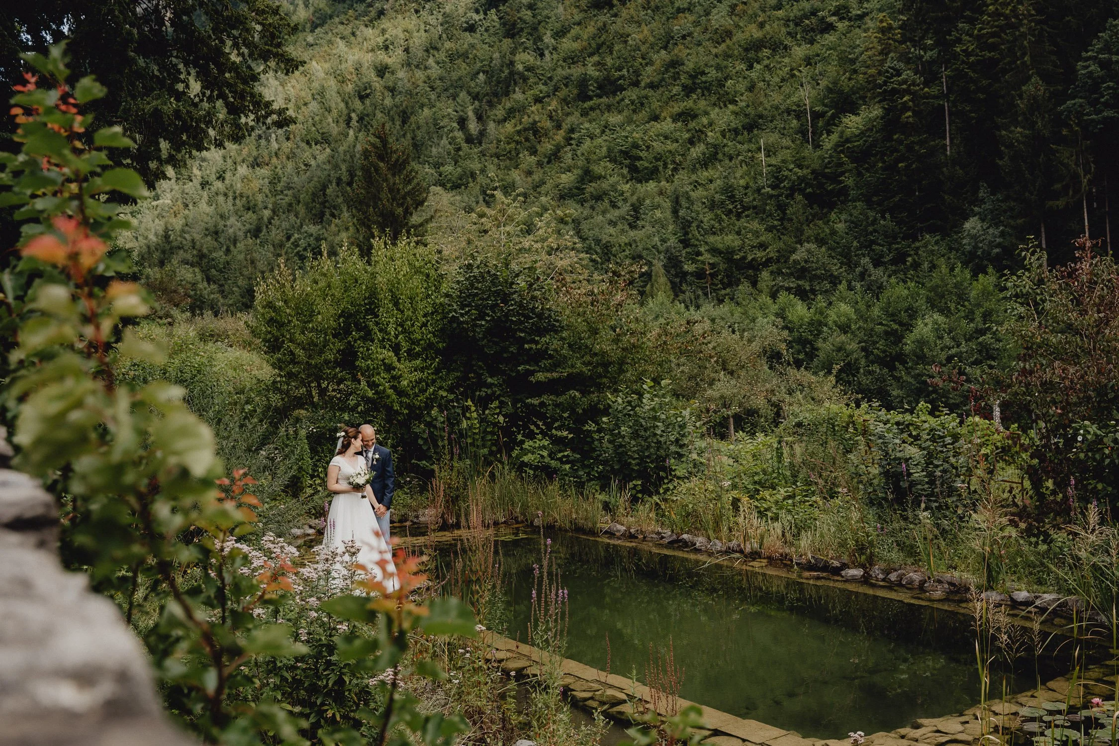 Ein Brautpaar im weißen Kleid und Anzug steht an einem kleinen Teich in einem grünen, bewaldeten Gebiet.
