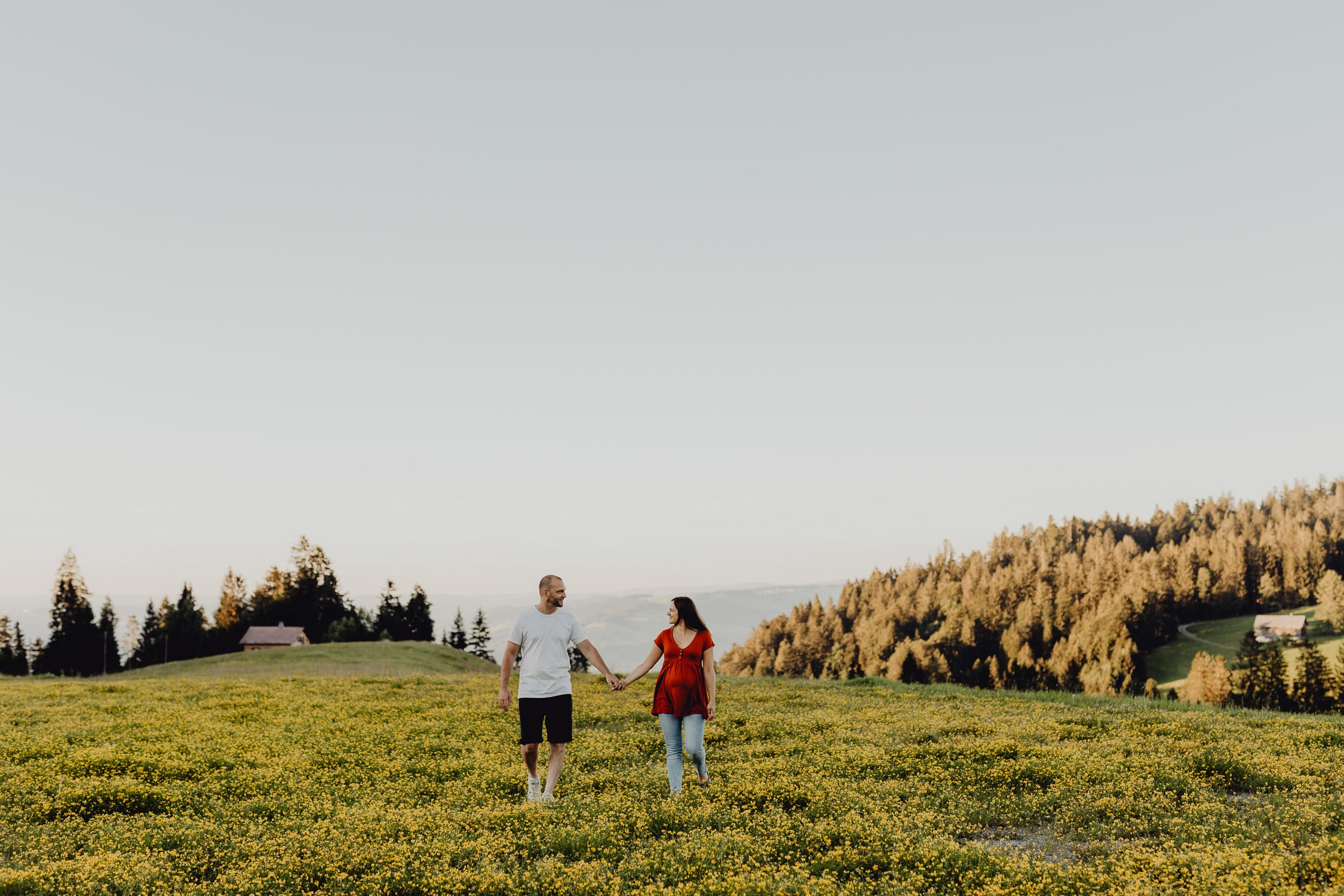 Ein Paar hält sich an einem sonnigen Tag in einer blühenden grünen Wiese mit gelben Blumen. Im Hintergrund befinden sich Bäume und Hügel.