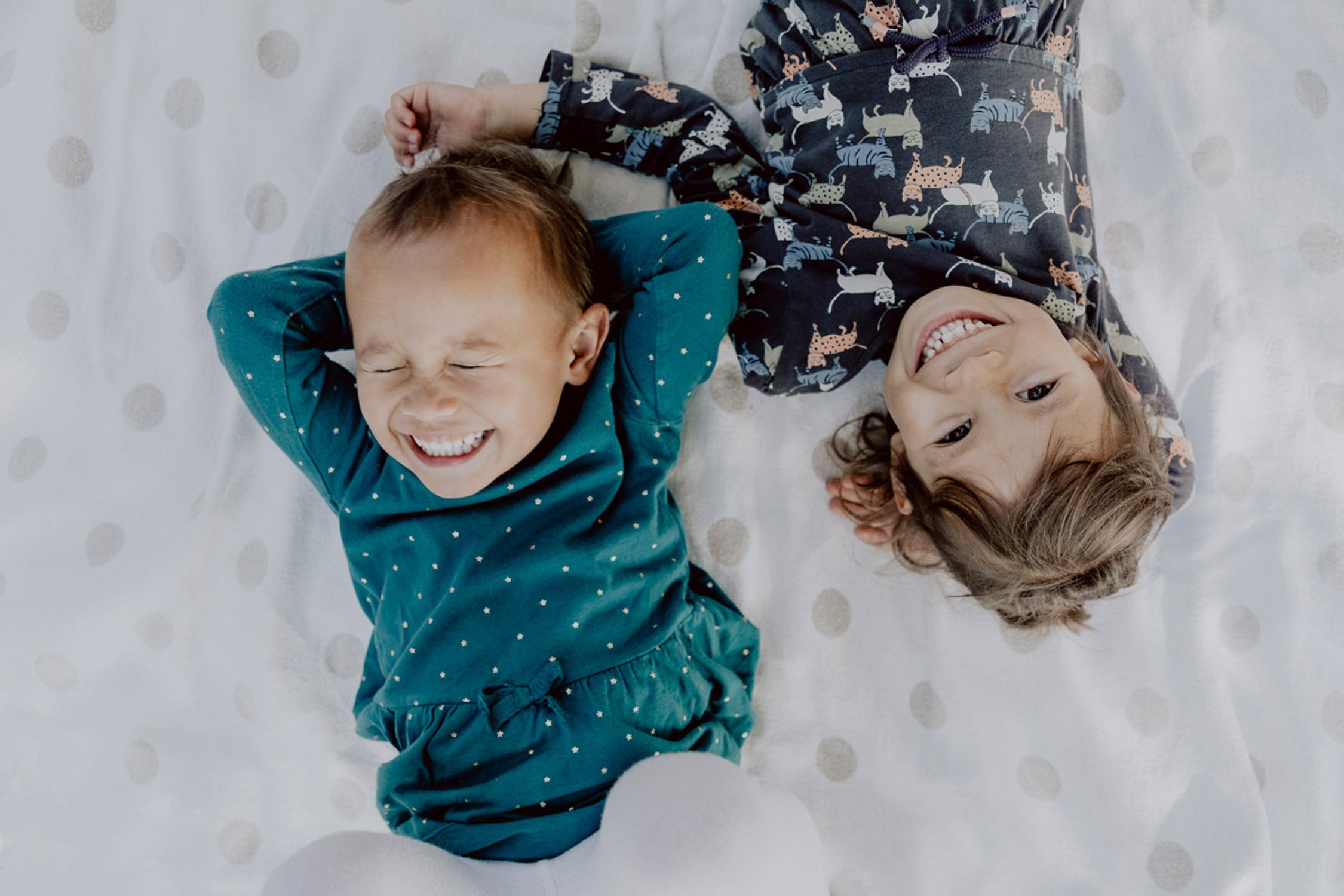 Zwei lachende Kinder, ein Junge und ein Mädchen, liegen auf einem weißen Bett mit beige Punkten. Der Junge trägt ein blaues Kleid mit weißen Sternen, das Mädchen ein dunkles Kleid mit Tiermustern.