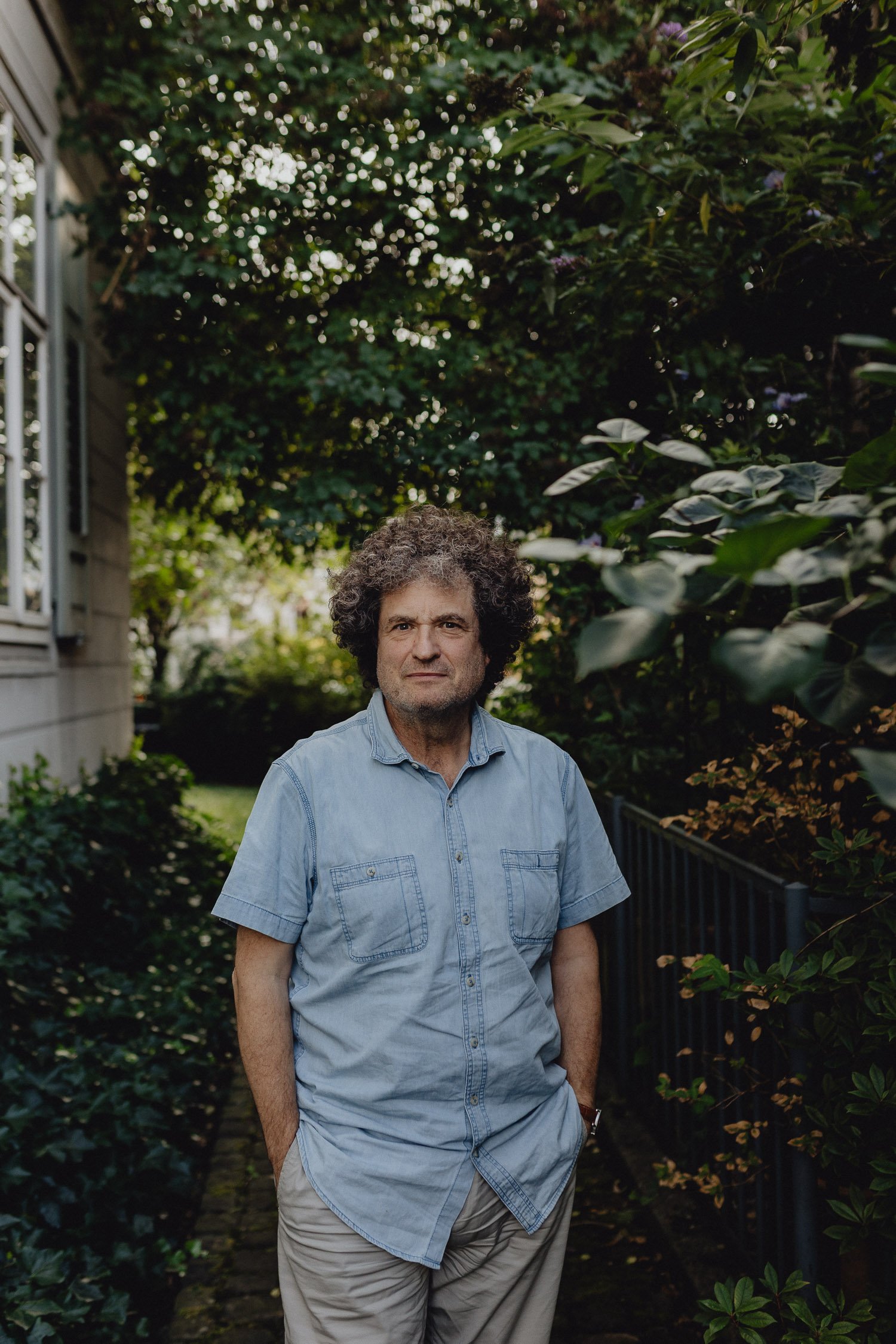 Ein Mann mit lockigem Haar trägt ein hellblaues Hemd und steht auf einem Gehweg in einem Garten mit vielen Büschen und Bäumen.