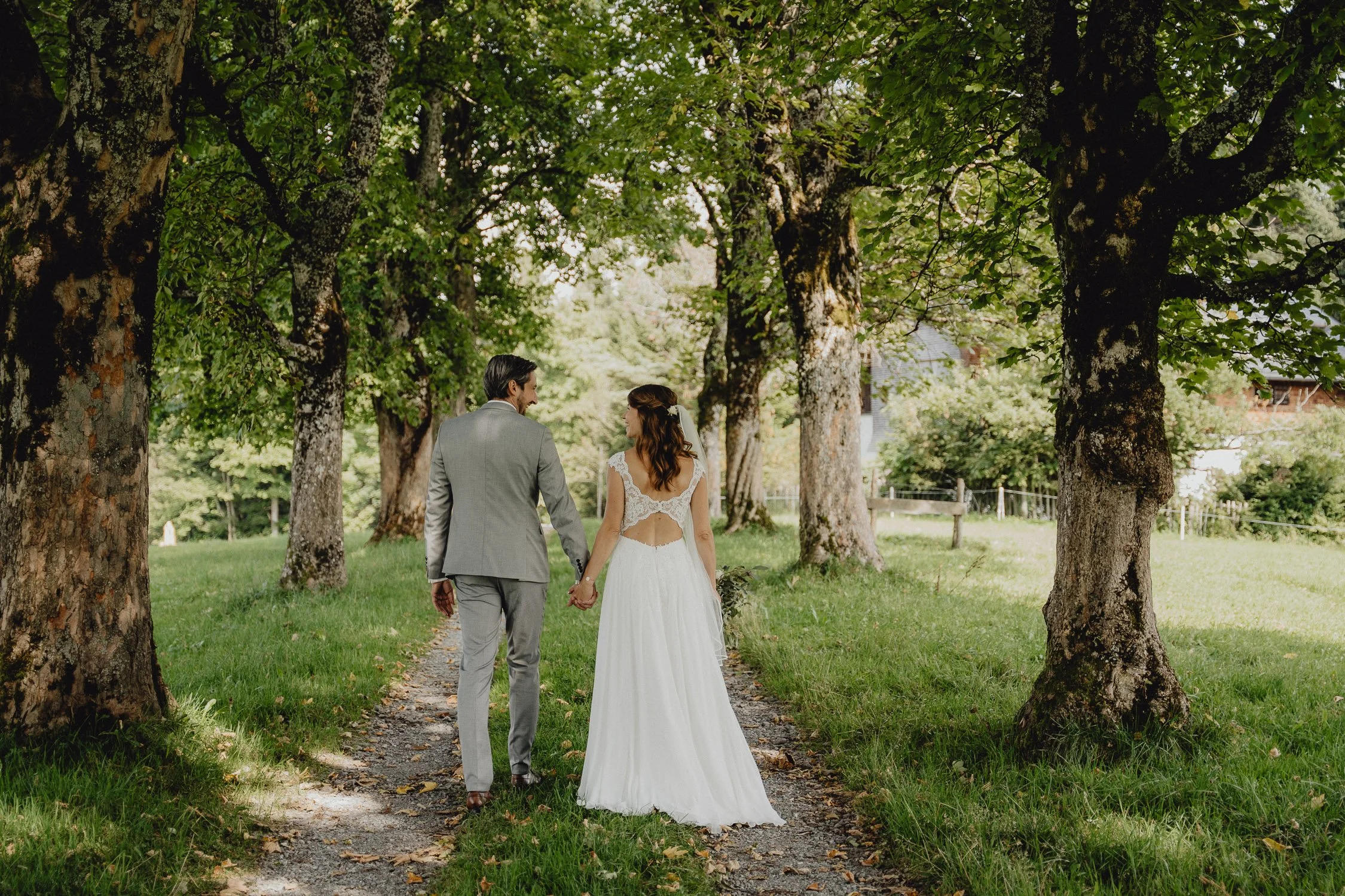 Ein Brautpaar, das Hand in Hand einen Baumweg entlang spaziert, umgeben von grünen Bäumen auf einer Wiese bei Sonnenlicht.