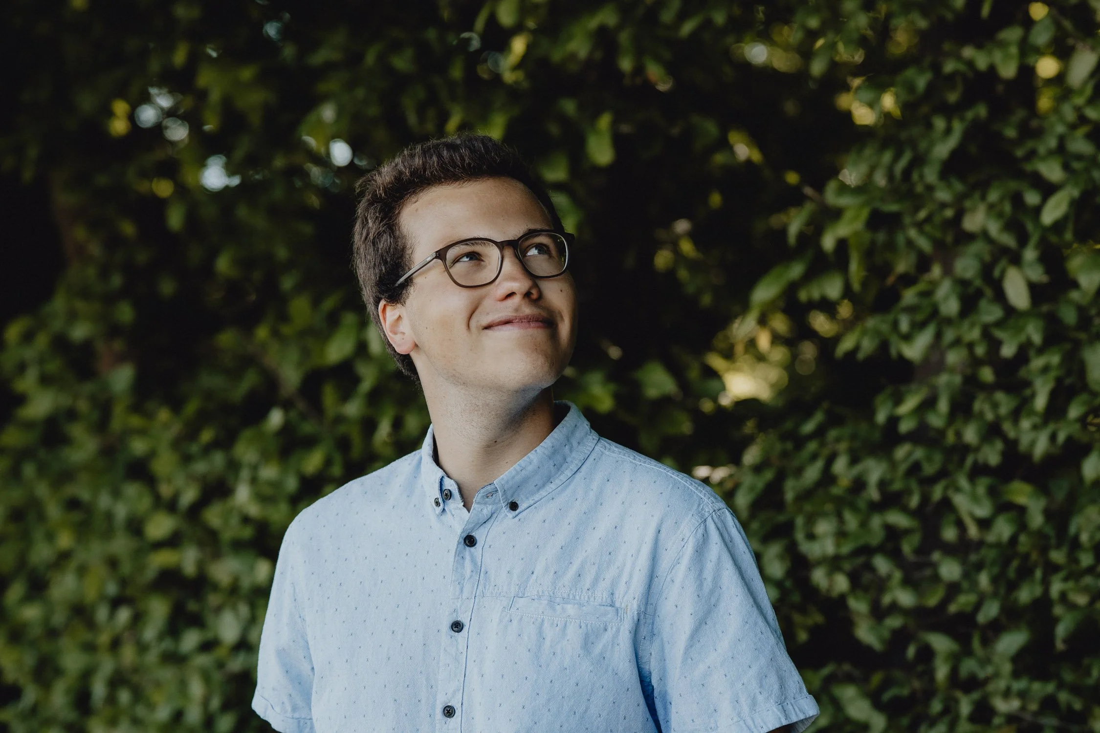 Junge Frau mit Brille steht vor einer grünen Hecke, lächelt leicht und schaut nach oben.