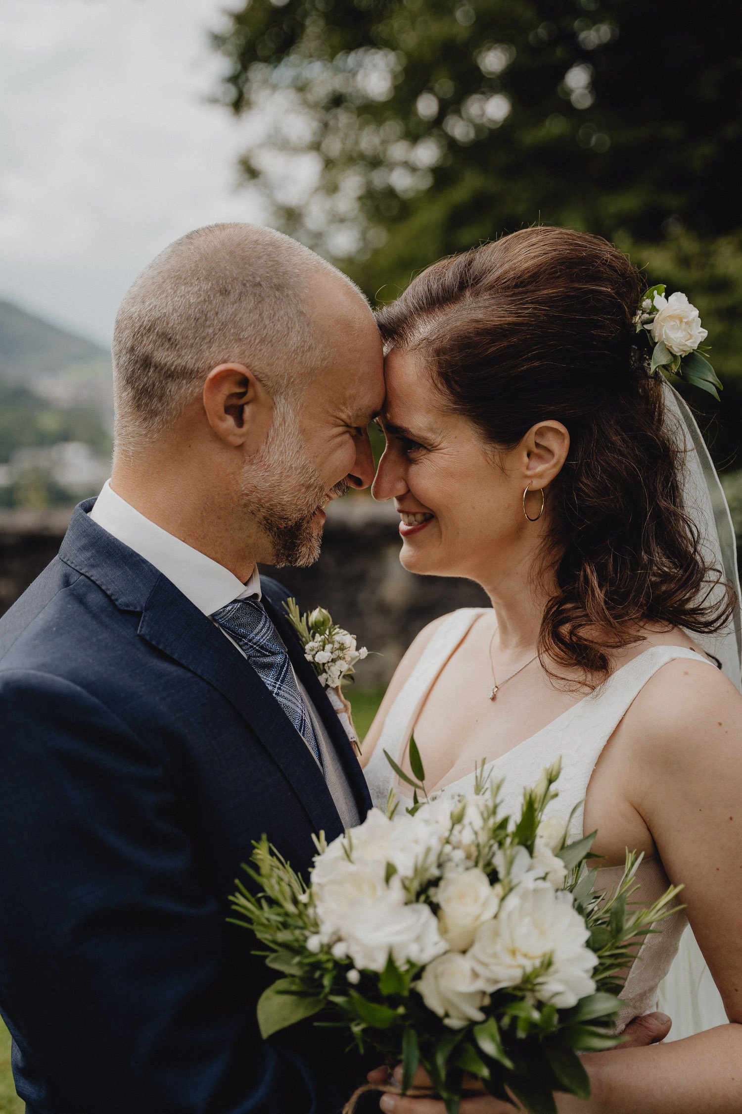 Ein Brautpaar steht sich bei ihrer Hochzeit gegenüber, ihre Stirn berühren sich, sie lachen und halten ein Blumenbouquet.