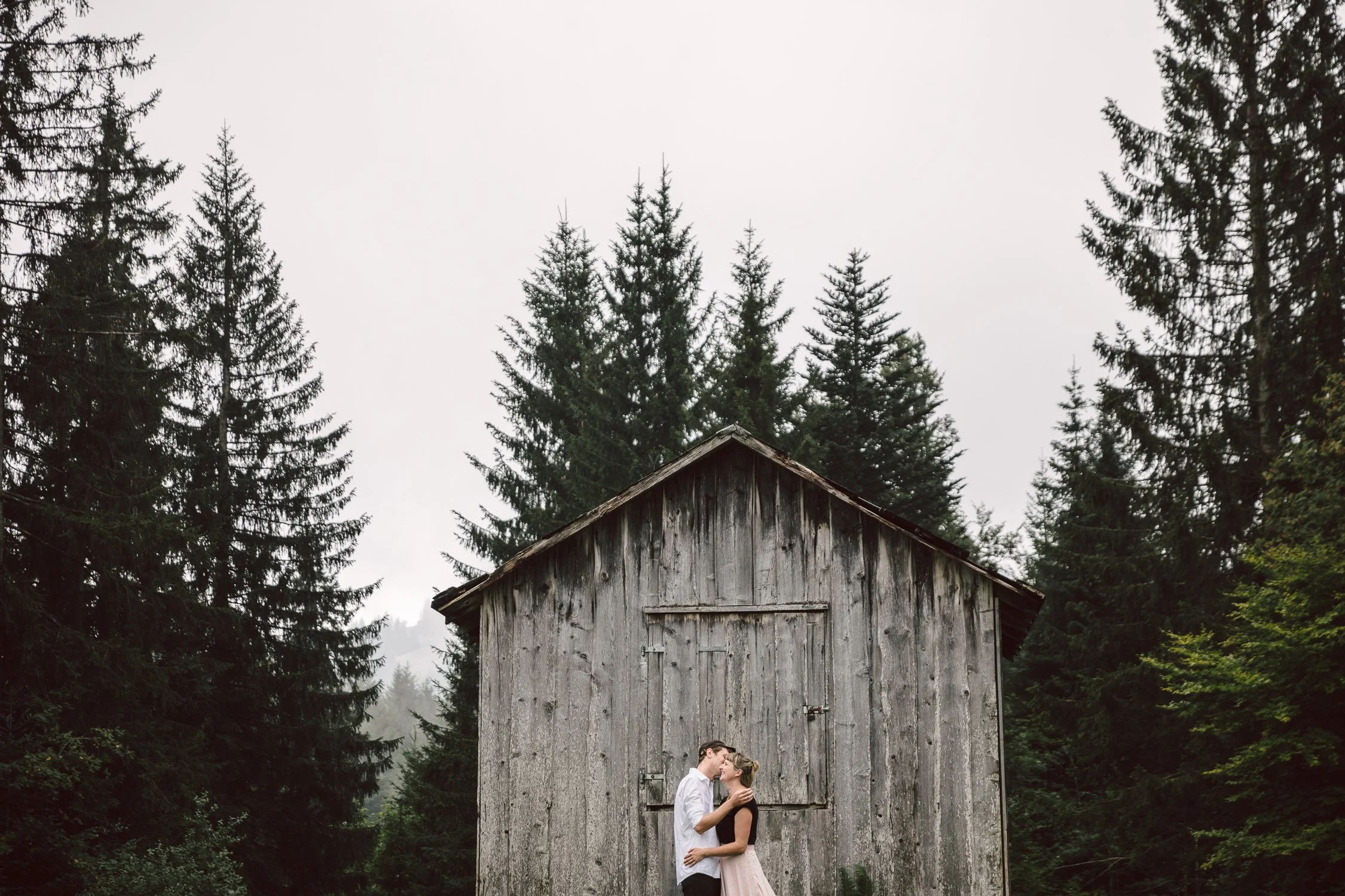 Ein verliebtes Paar küsst sich vor einer alten Holzhütte, umgeben von Tannenbäumen.