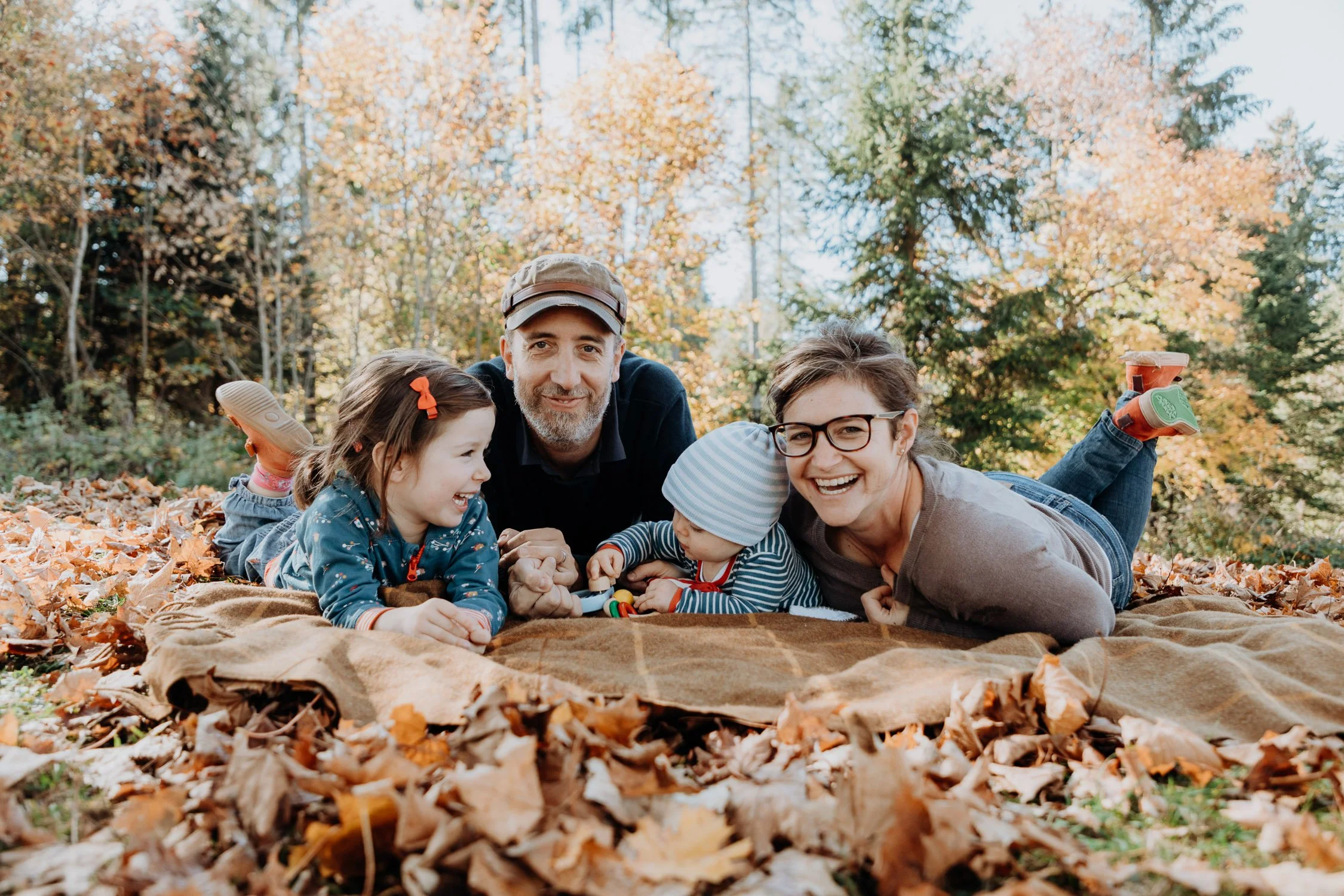 Familie mit zwei Erwachsenen und zwei Kindern liegt auf dem Boden in einem Herbstwald, umgeben von fallenden Blättern, lachend und spielend.