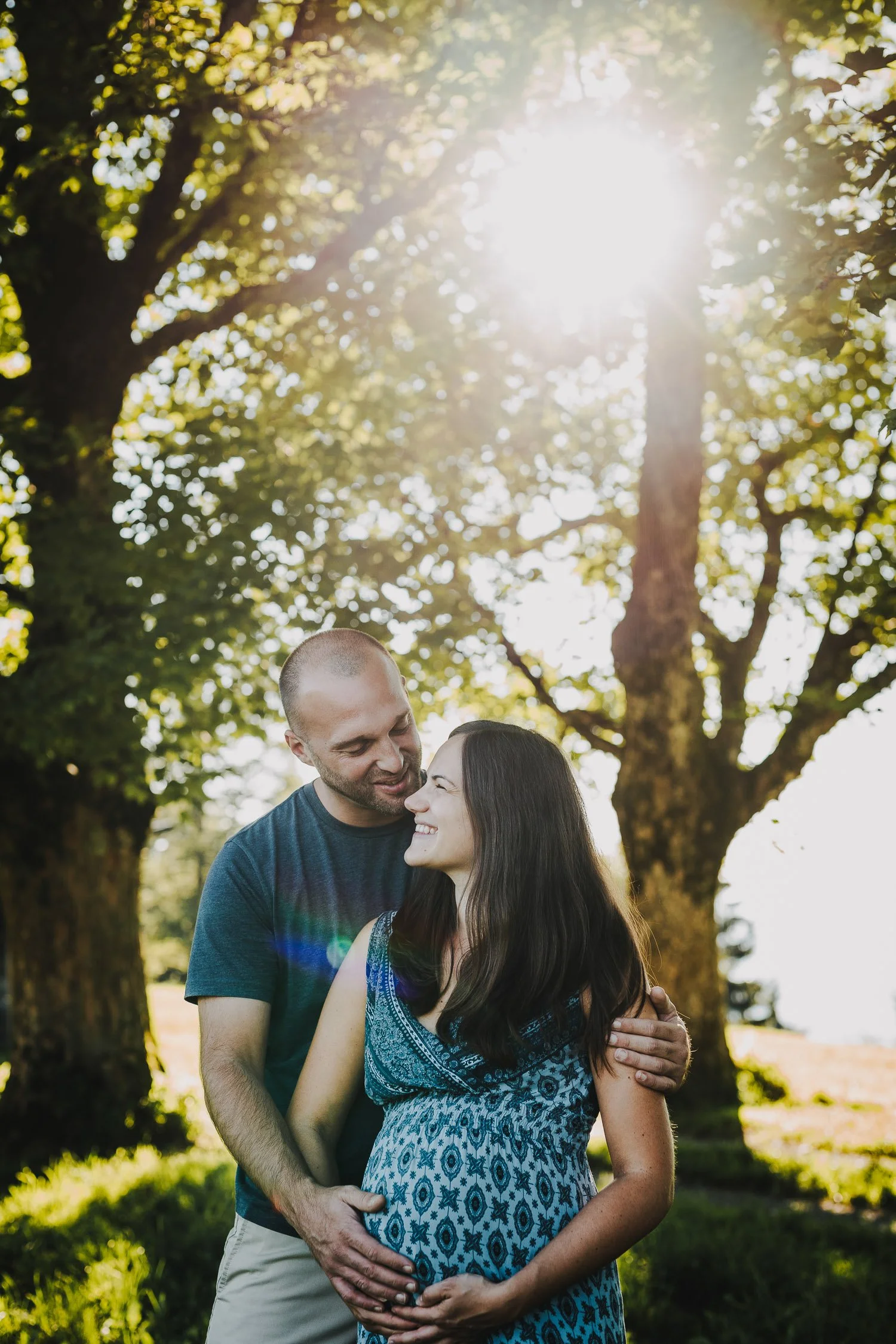 Ein glückliches Paar steht im Freien vor großen Bäumen, die Sonnenstrahlen durch die Blätter schicken. Die schwangere Frau trägt ein blaues Kleid mit Mustern, der Mann umarmt sie liebevoll und beide lächeln sich an.