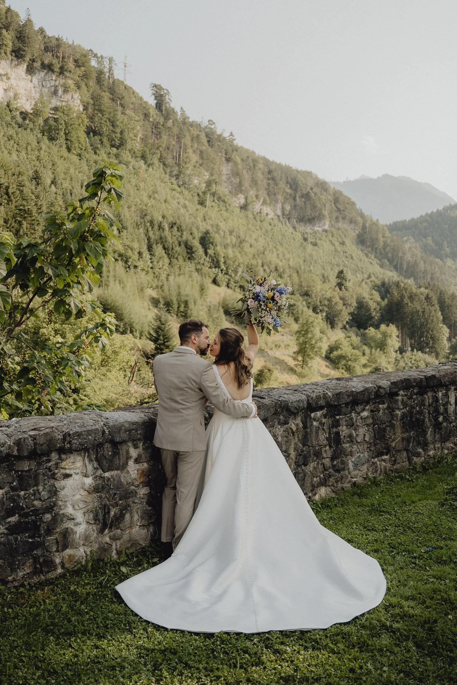 Ein Brautpaar steht an einer Steinmauer vor einer bergigen, bewaldeten Landschaft an einem Tag mit bewölktem Himmel. Die Braut trägt ein weißes Brautkleid und hält einen Blumenstrauß hoch, während der Bräutigam sie umarmt.