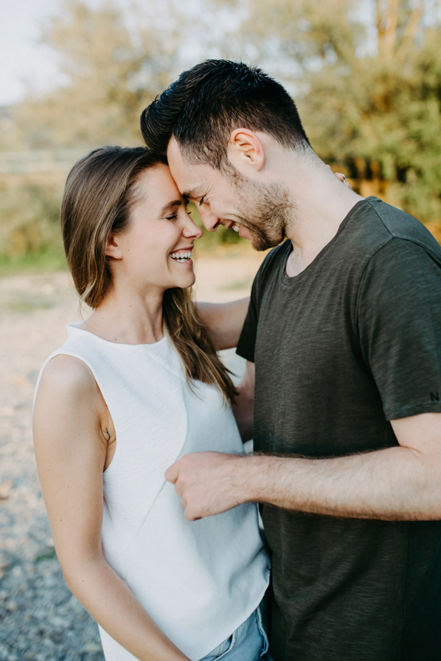 Paar lacht sich draußen in der Natur mit Kopf an Kopf, die Frau trägt ein weißes Hemd, der Mann ein dunkles T-Shirt.