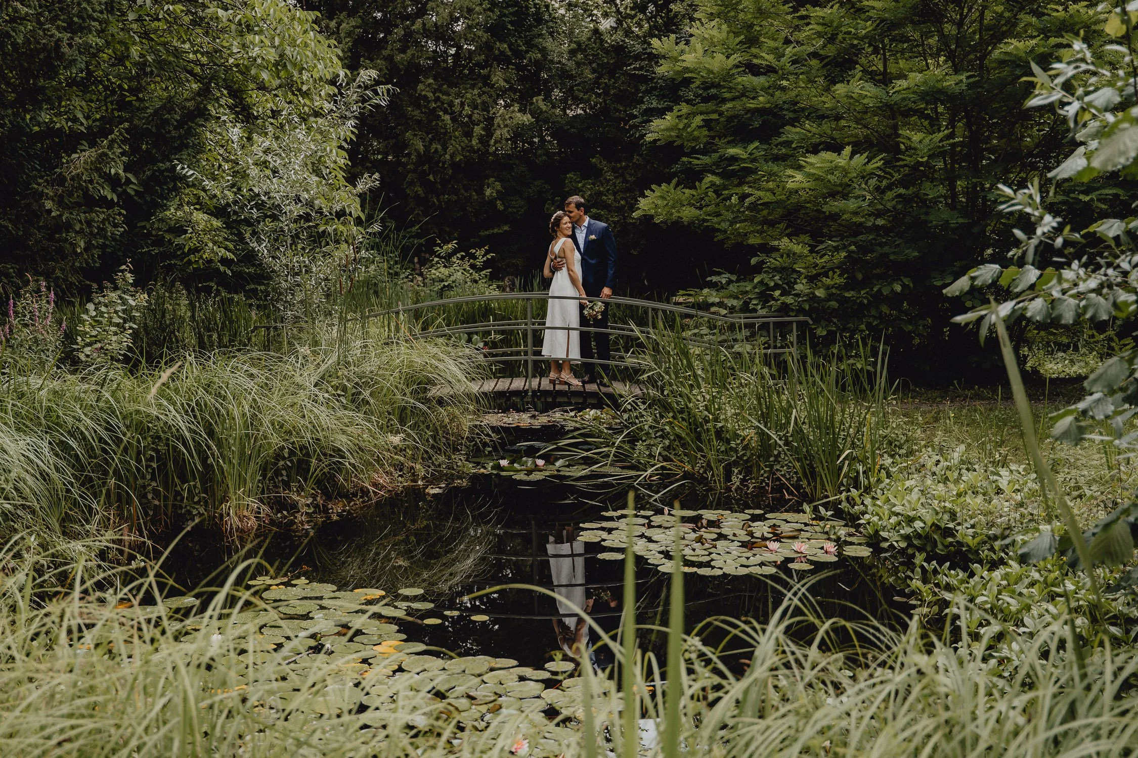 Ein Brautpaar steht auf einer kleinen Brücke über einem Teich mit Seerosen in einem grünen, bewaldeten Garten.