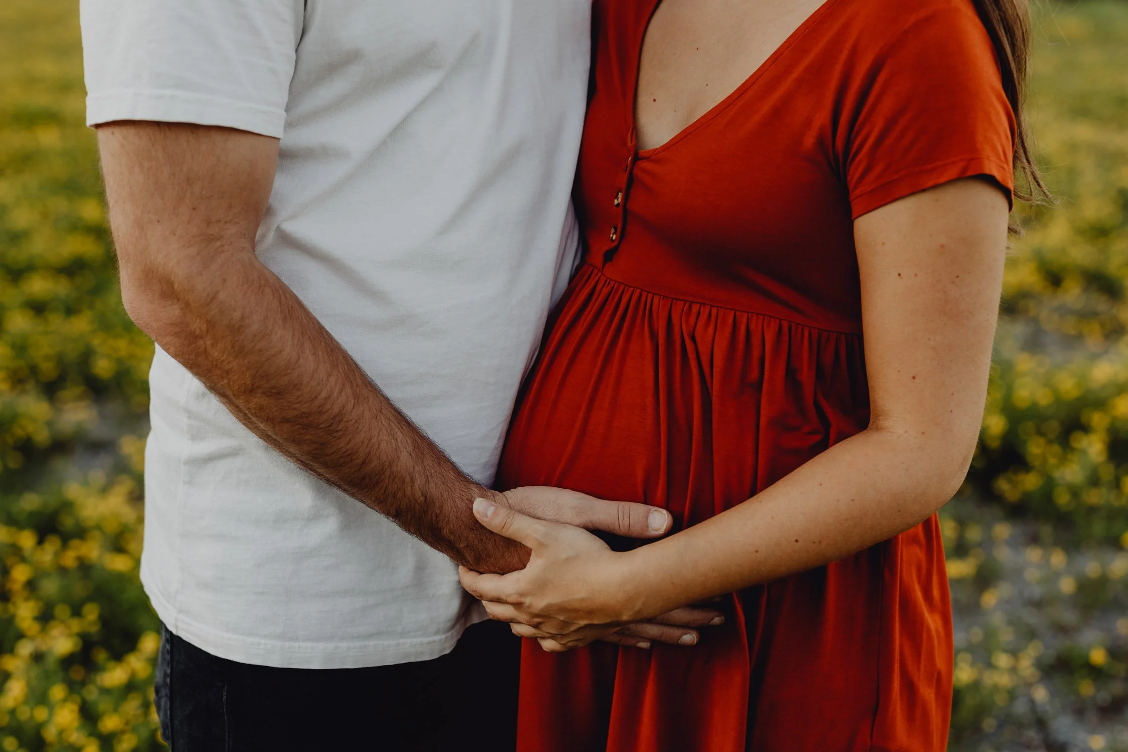 Ein schwangere Frau in einem roten Kleid hält den Bauch eines Mannes in einem weißen T-Shirt bei einem Spaziergang im Freien fest.