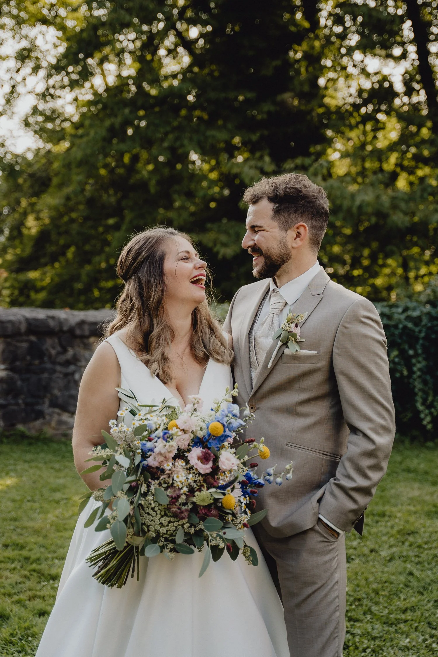 Ein glückliches Brautpaar bei einer Hochzeit im Freien, die Braut hält einen bunten Blumenstrauß, beide lachen sich an, im Hintergrund Bäume.