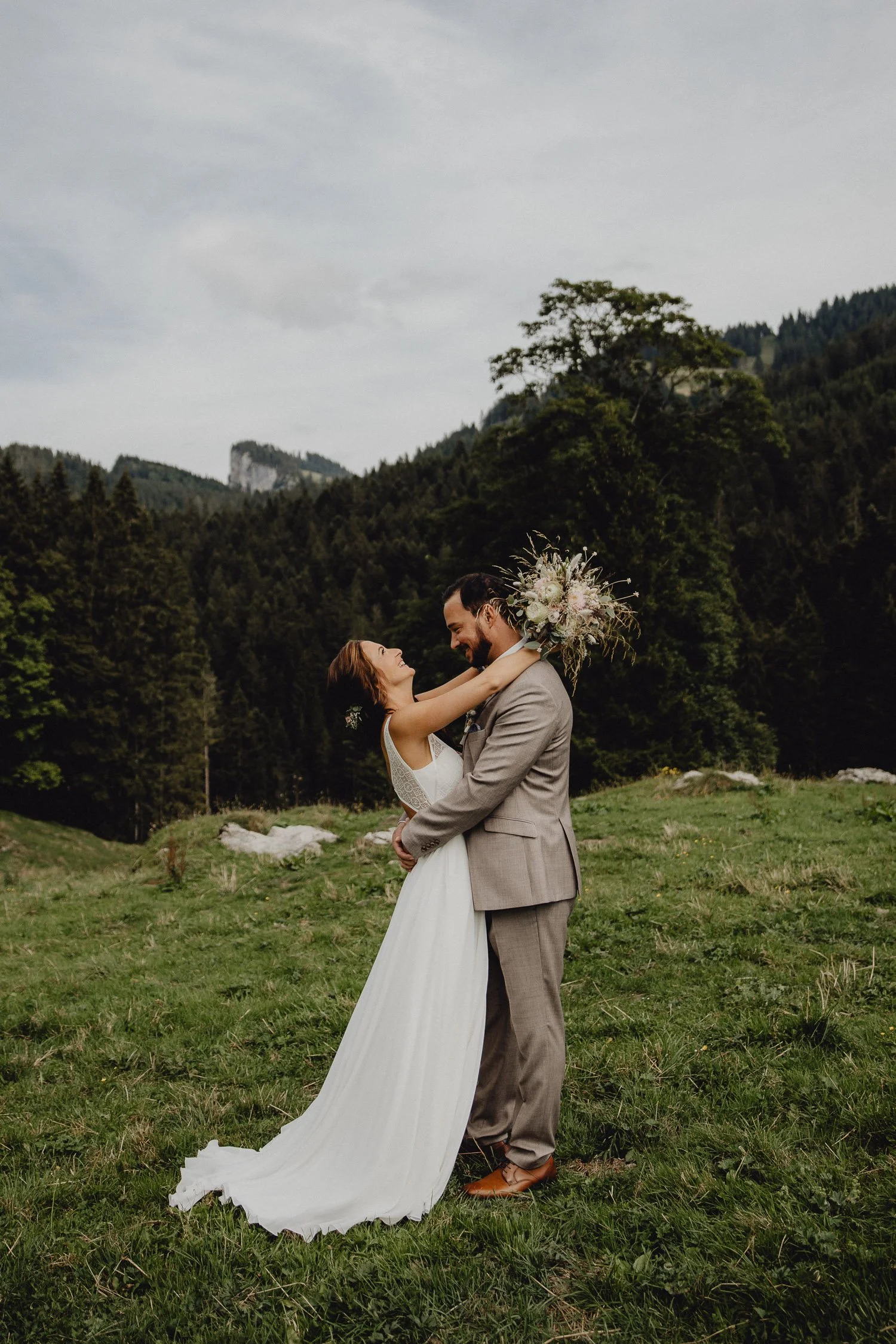Braut und Bräutigam umarmen sich auf einer Wiese in den Bergen, umgeben von Bäumen, bei Sonnen- oder Wolkenhimmel, outdoor Hochzeitsfotografie