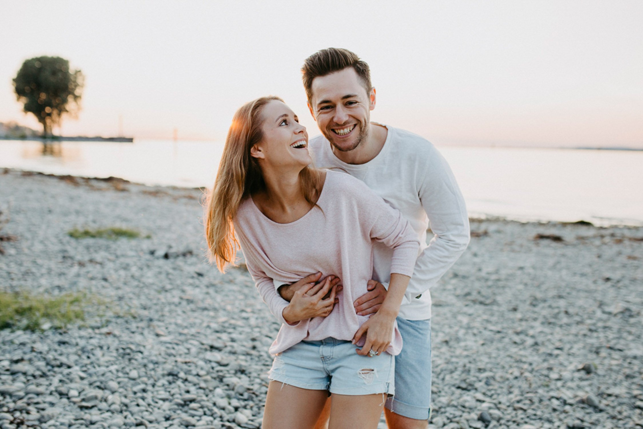 Ein lachendes Paar steht am Strand bei Sonnenuntergang, die Frau trägt ein pinkfarbenes Top und Shorts, der Mann trägt ein weißes Oberteil und Shorts, sie scheinen Spaß zu haben.