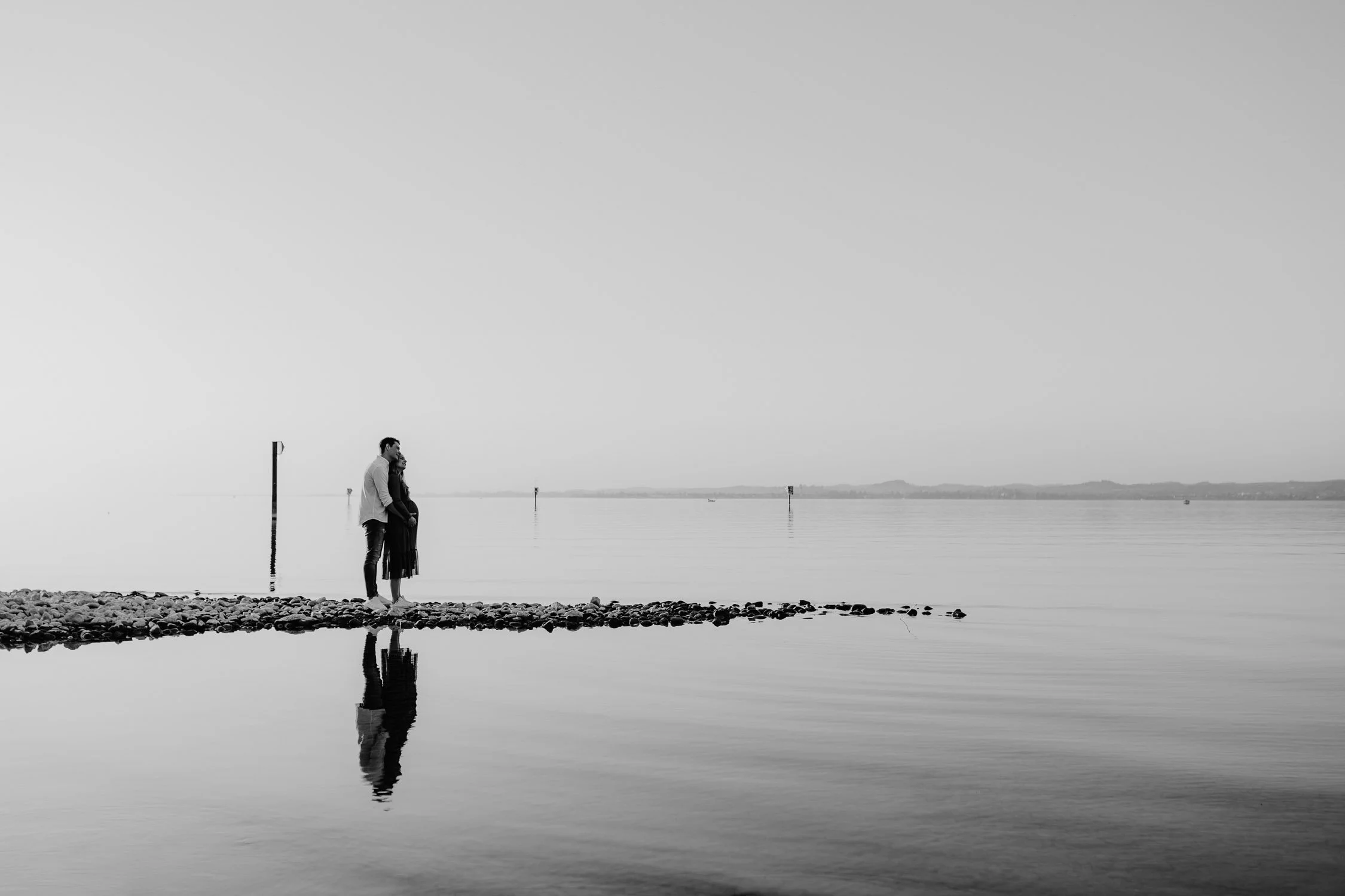 Ein Pärchen steht am Ufer eines ruhigen Sees, ihre Reflexion ist im Wasser sichtbar, im Hintergrund sind Markierungen im Wasser und entfernte Hügel zu sehen.