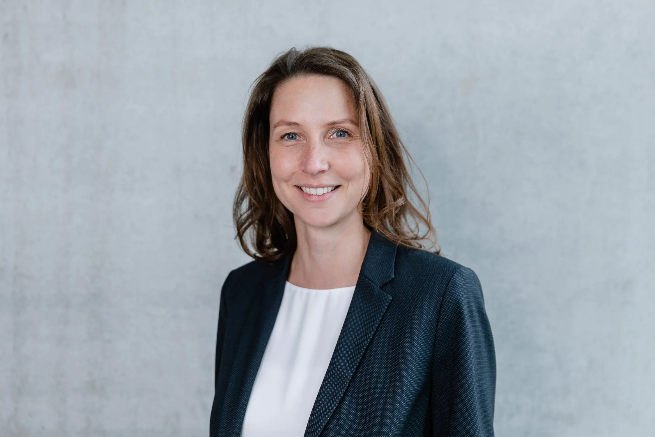 Porträt einer lächelnden Frau im Business-Outfit vor einem grauen Hintergrund.