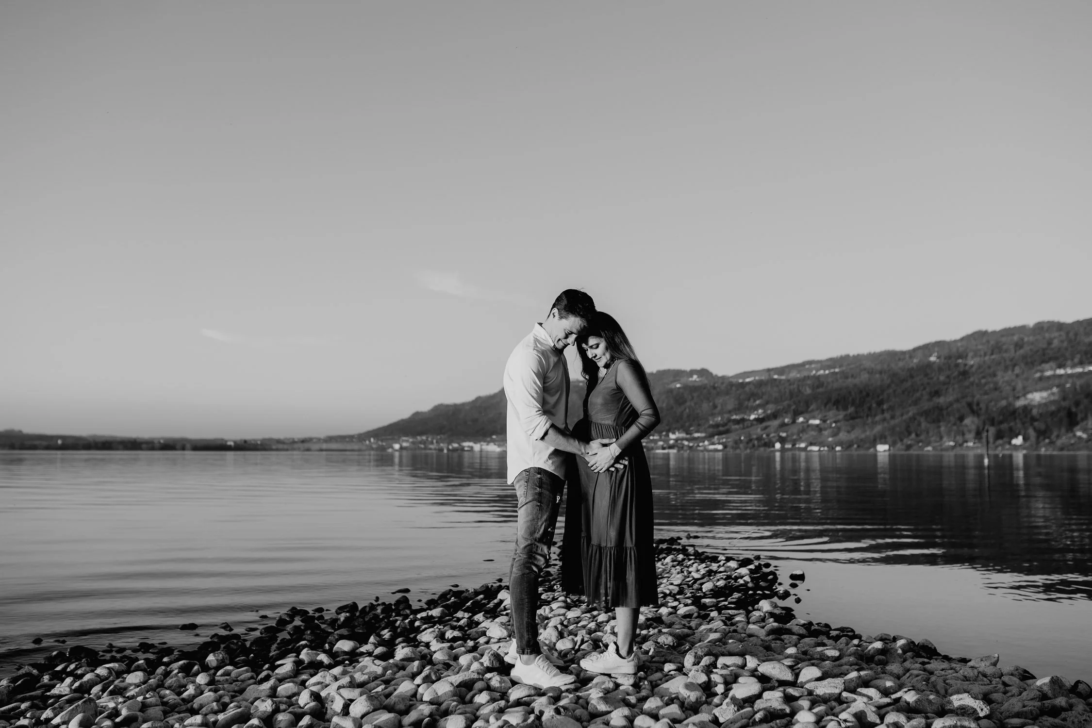Ein Paar steht Händchen haltend auf einem Steg am Wasser, lächelnd, bei Sonnenuntergang, im Schwarz-Weiß-Foto.