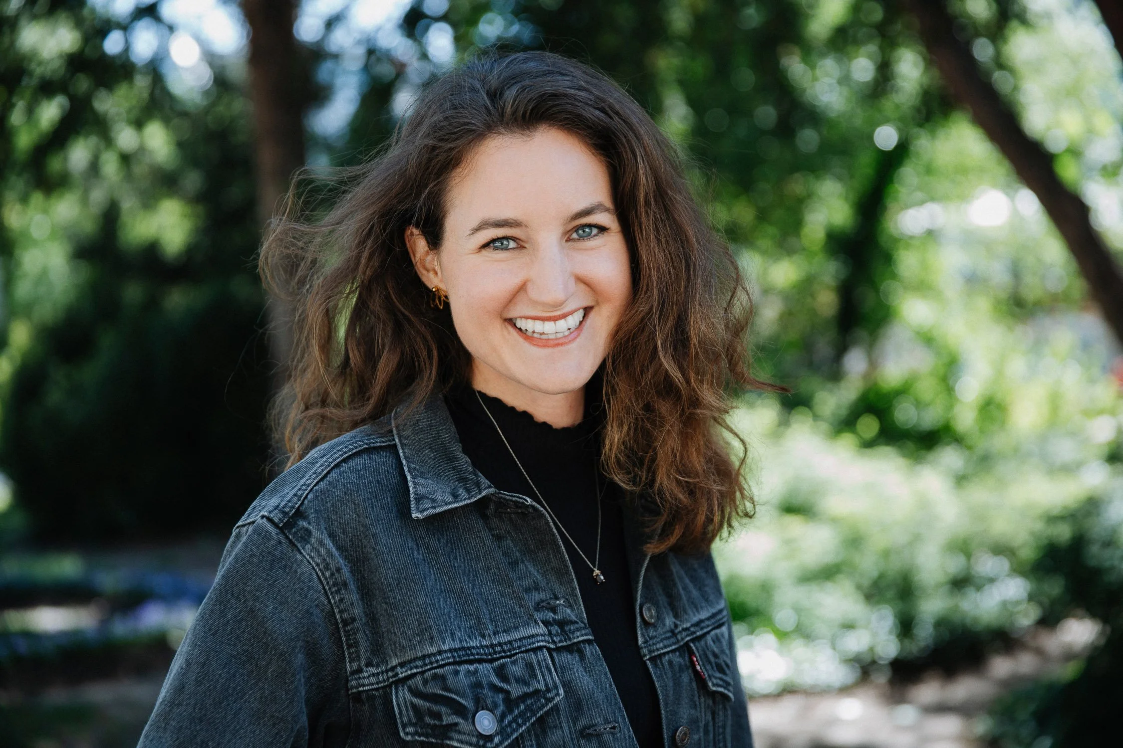 Eine lachende Frau mit braunen, lockigen Haaren und blauen Augen, trägt eine dunkle Jeansjacke und ein schwarzes Oberteil, steht vor einem grünen, bewaldeten Hintergrund.