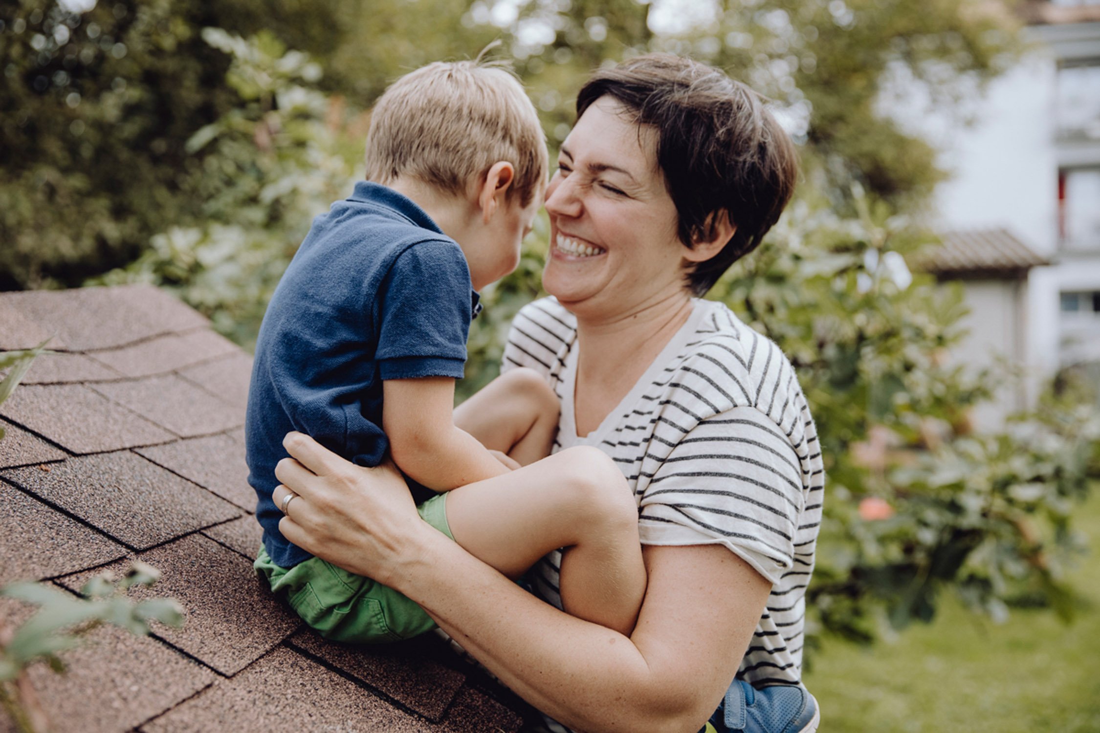 Eine Frau hält lachenden Jungen auf einem Dach, umgeben von Bäumen und Häusern im Hintergrund.