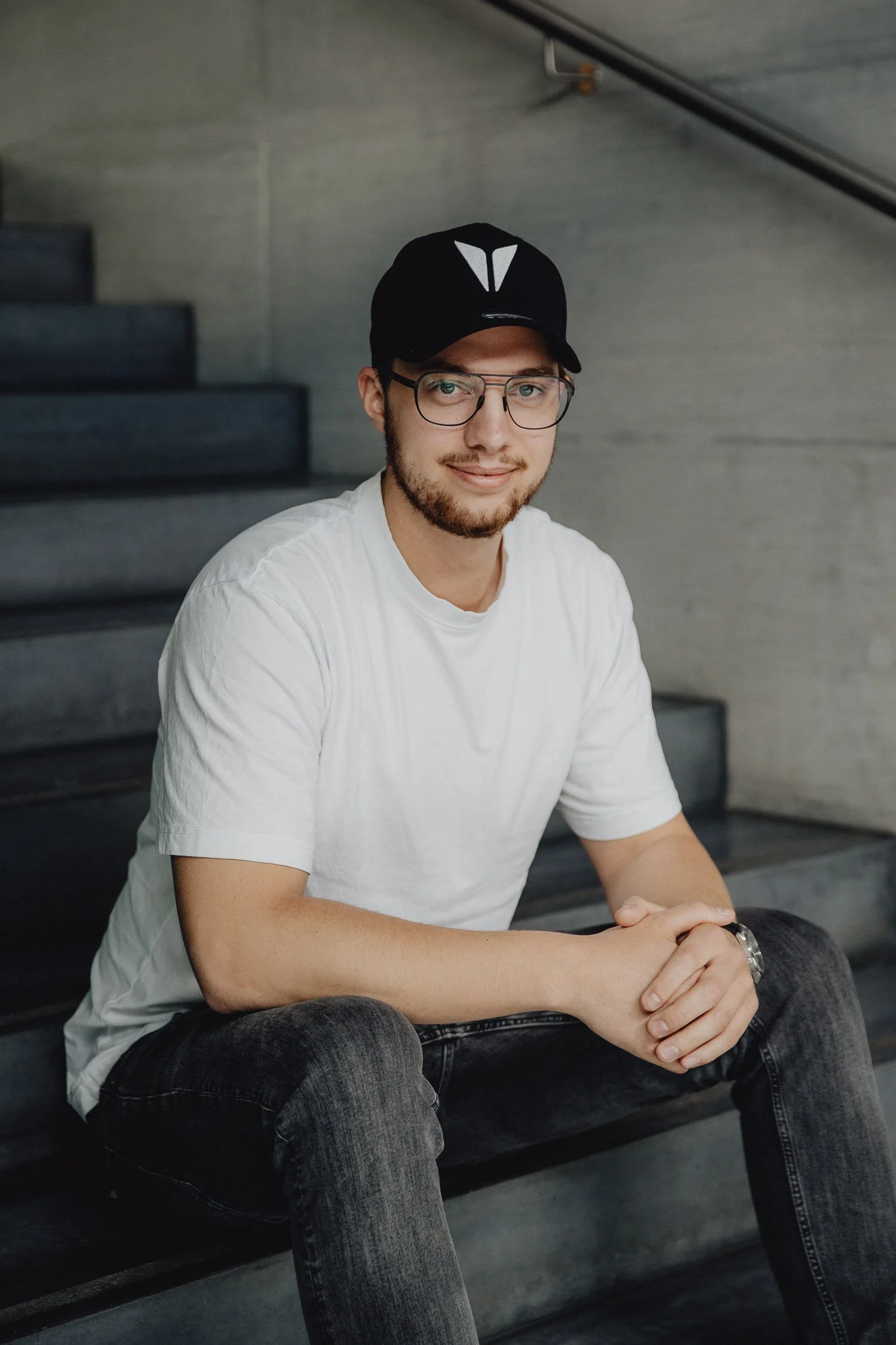 Junger Mann sitzend auf dunklen Stufen, trägt weißes T-Shirt, schwarze Jeans, schwarzer Cap und Brille, lächelt leicht.