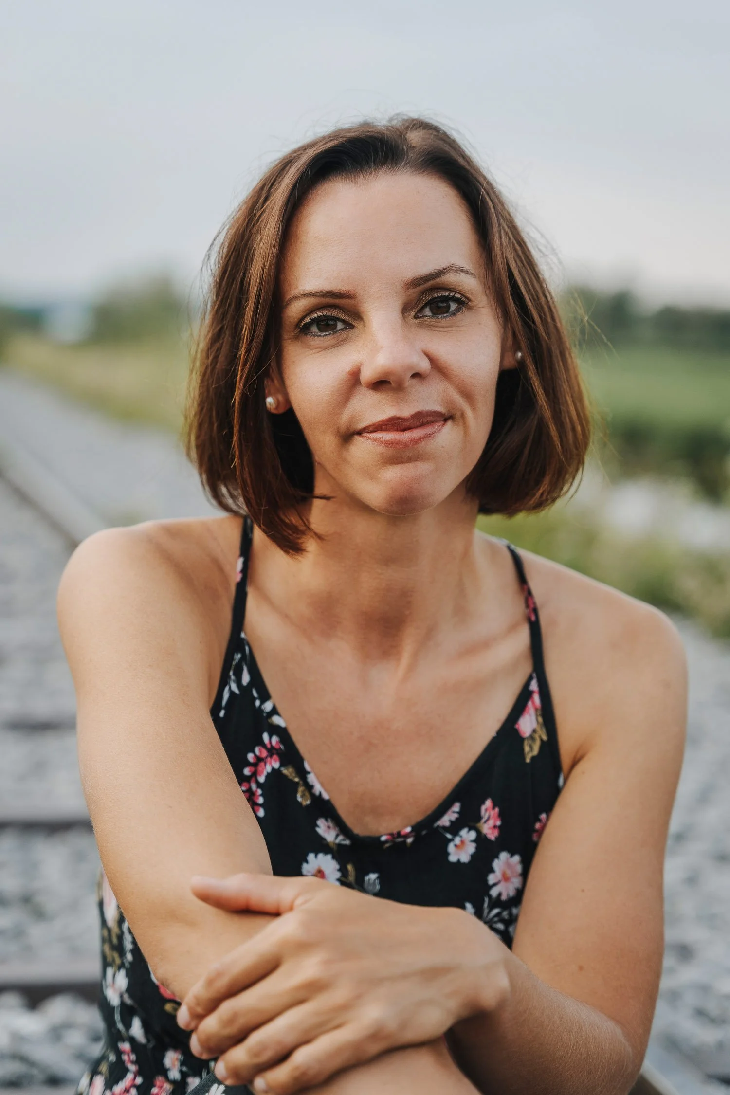 Nahaufnahme einer lächelnden Frau mit braunen Haaren, in einem schwarzen Sommerkleid mit Blumenmuster, auf einer Bahnstrecke im Freien.