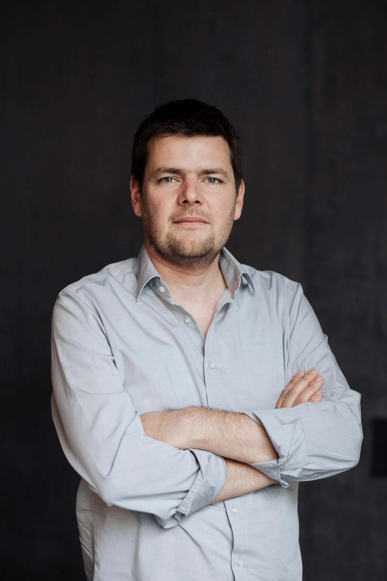 Porträt eines Mannes, der ein grau Hemd trägt, vor dunklem Hintergrund, Arme verschränkt