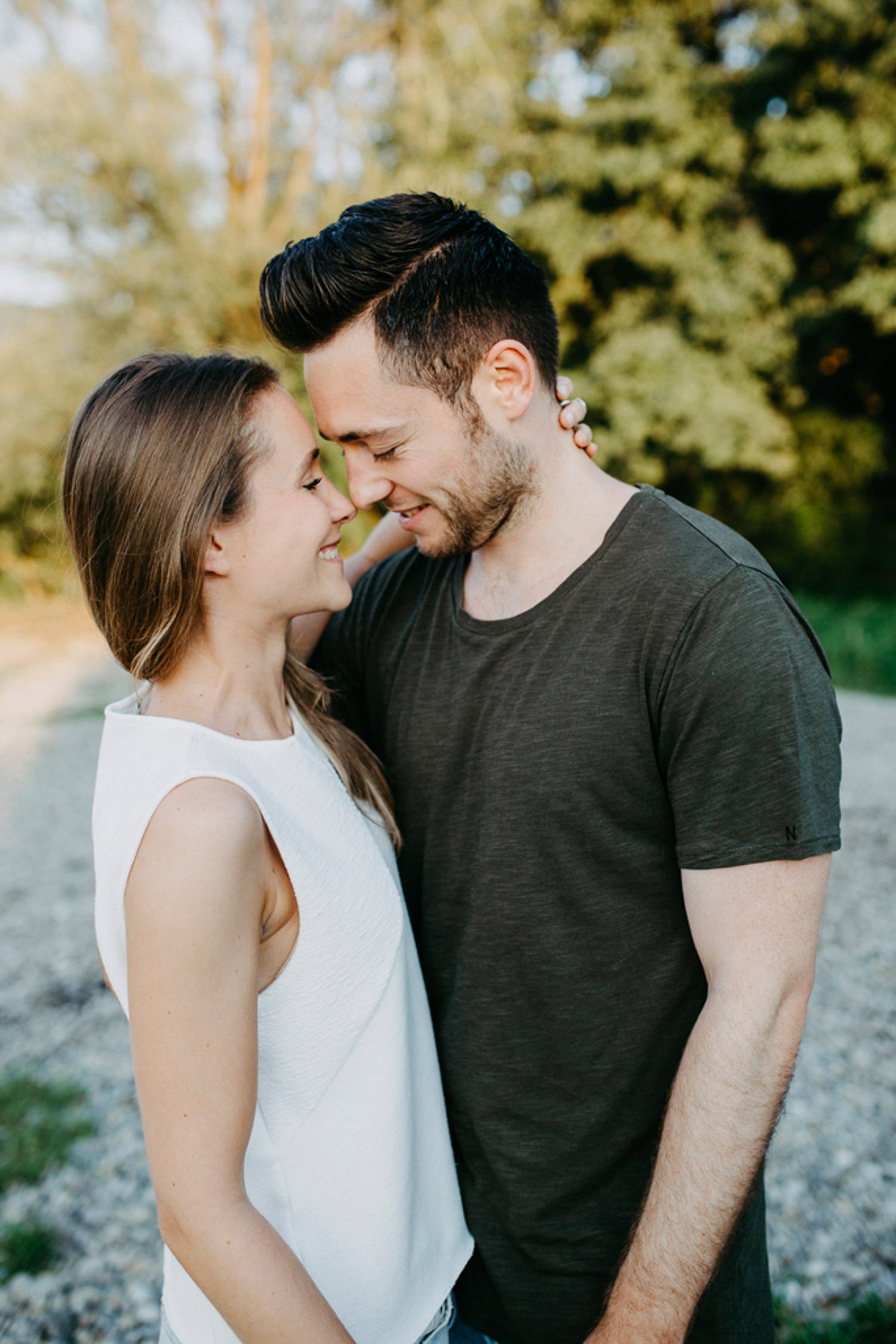 Ein Paar steht im Freien, sie lächeln sich an und berühren ihre Stirn, umgeben von Bäumen im Hintergrund.