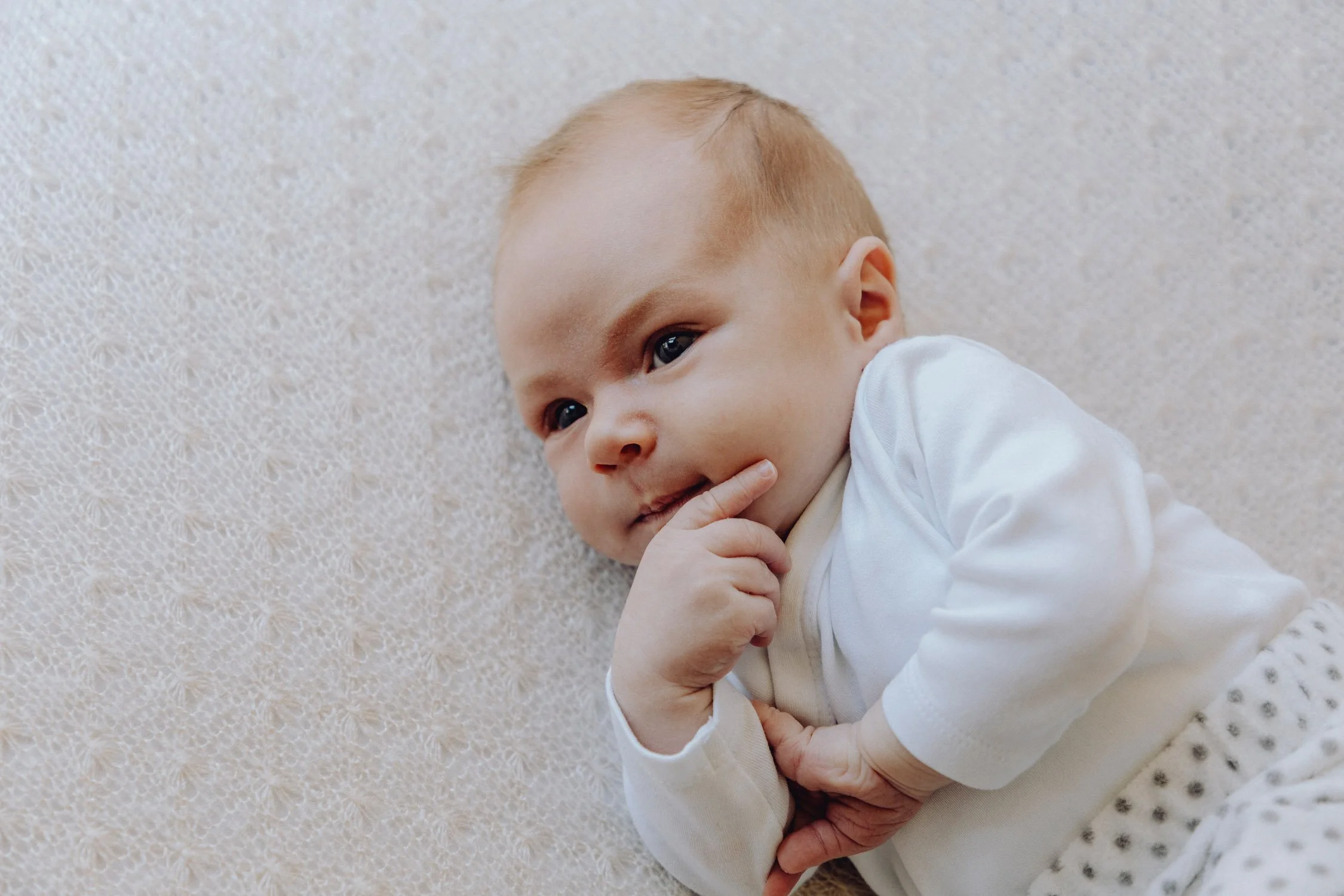 Ein Baby liegt auf einem hellen, strukturierten Teppich und berührt nachdenklich sein Kinn.