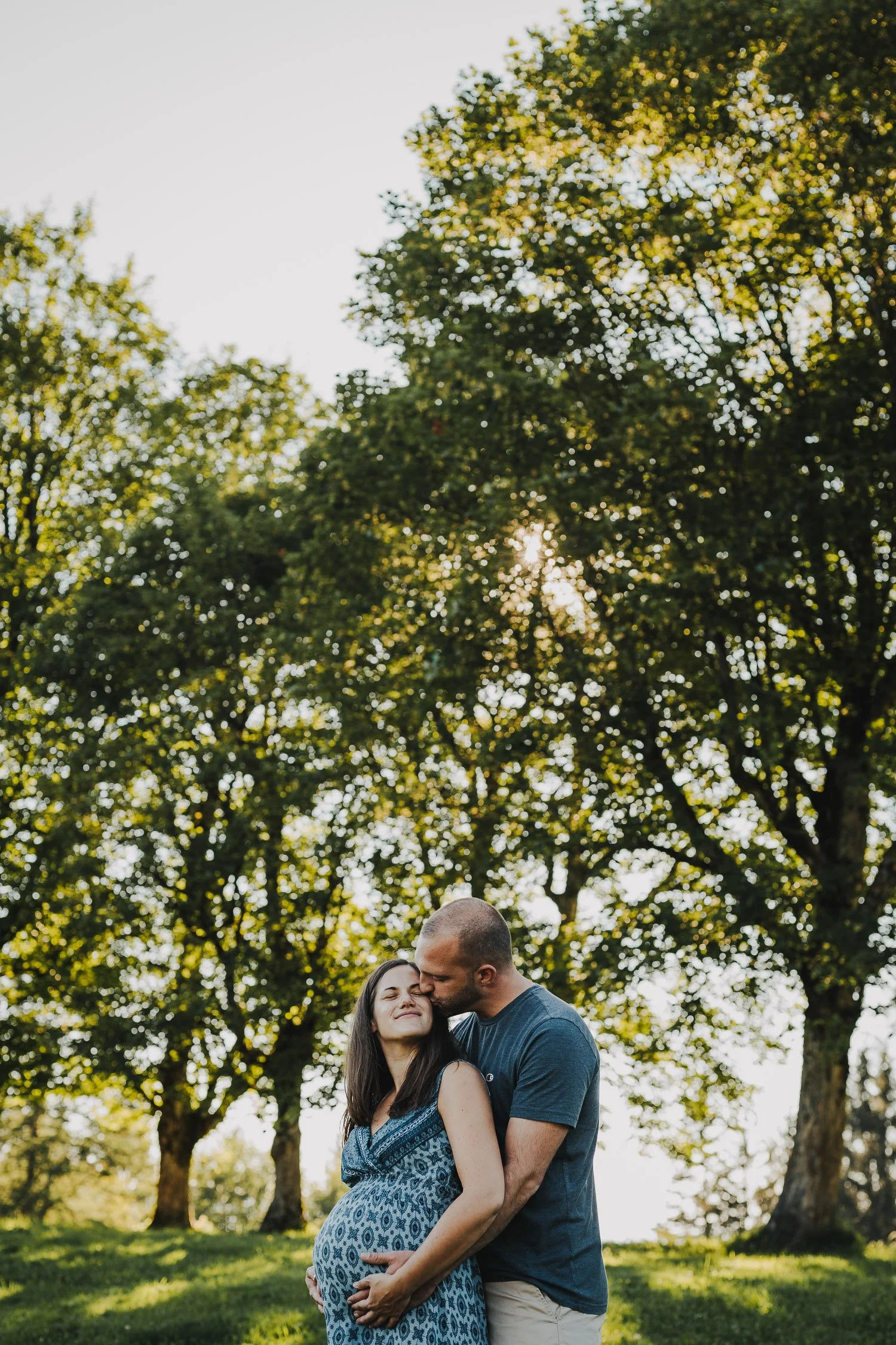 Ein schwangeres Paar umarmt sich liebevoll bei Sonnenuntergang, im Hintergrund große grüne Bäume.