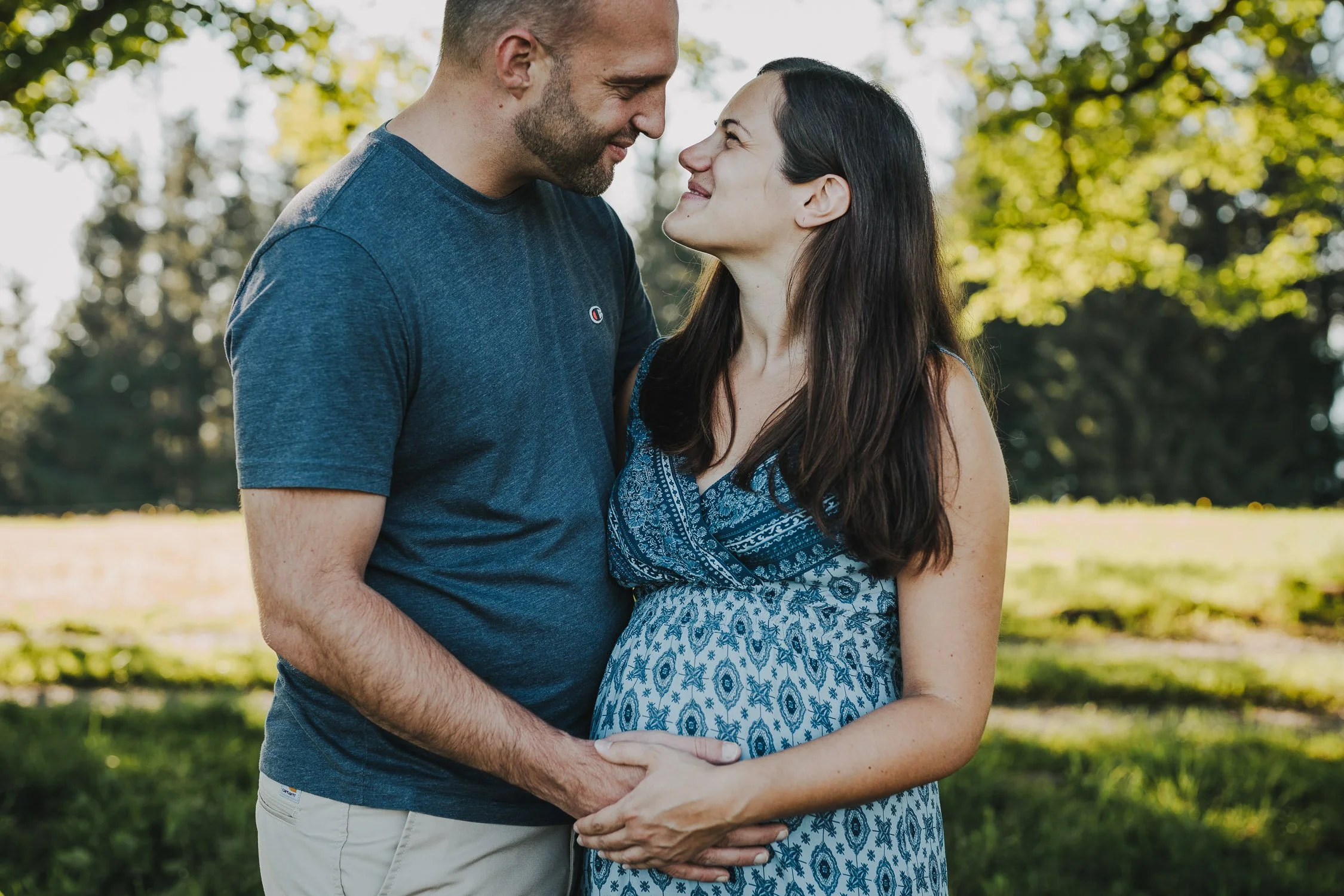 Ein schwangertes Paar steht lachend im Park, hält sich an den Händen und blickt sich liebevoll an.