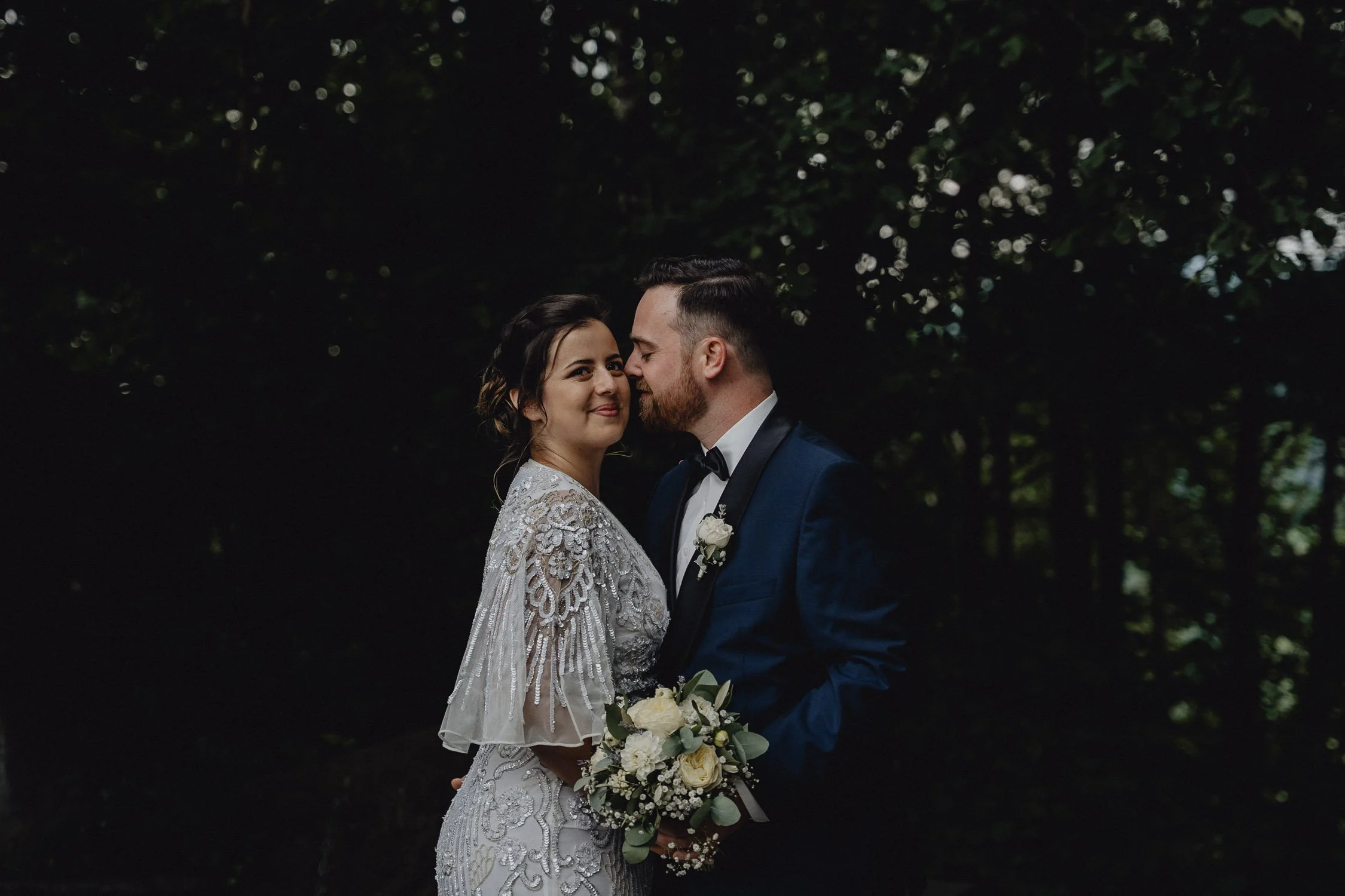 Hochzeitsfoto eines Paares im dunklen Wald, der Braut in einem weißen, verzierten Kleid, der Bräutigam in einem blauen Anzug mit Fliege, das Brautpaar steht sich nah gegenüber, die Braut hält einen Blumenstrauß.