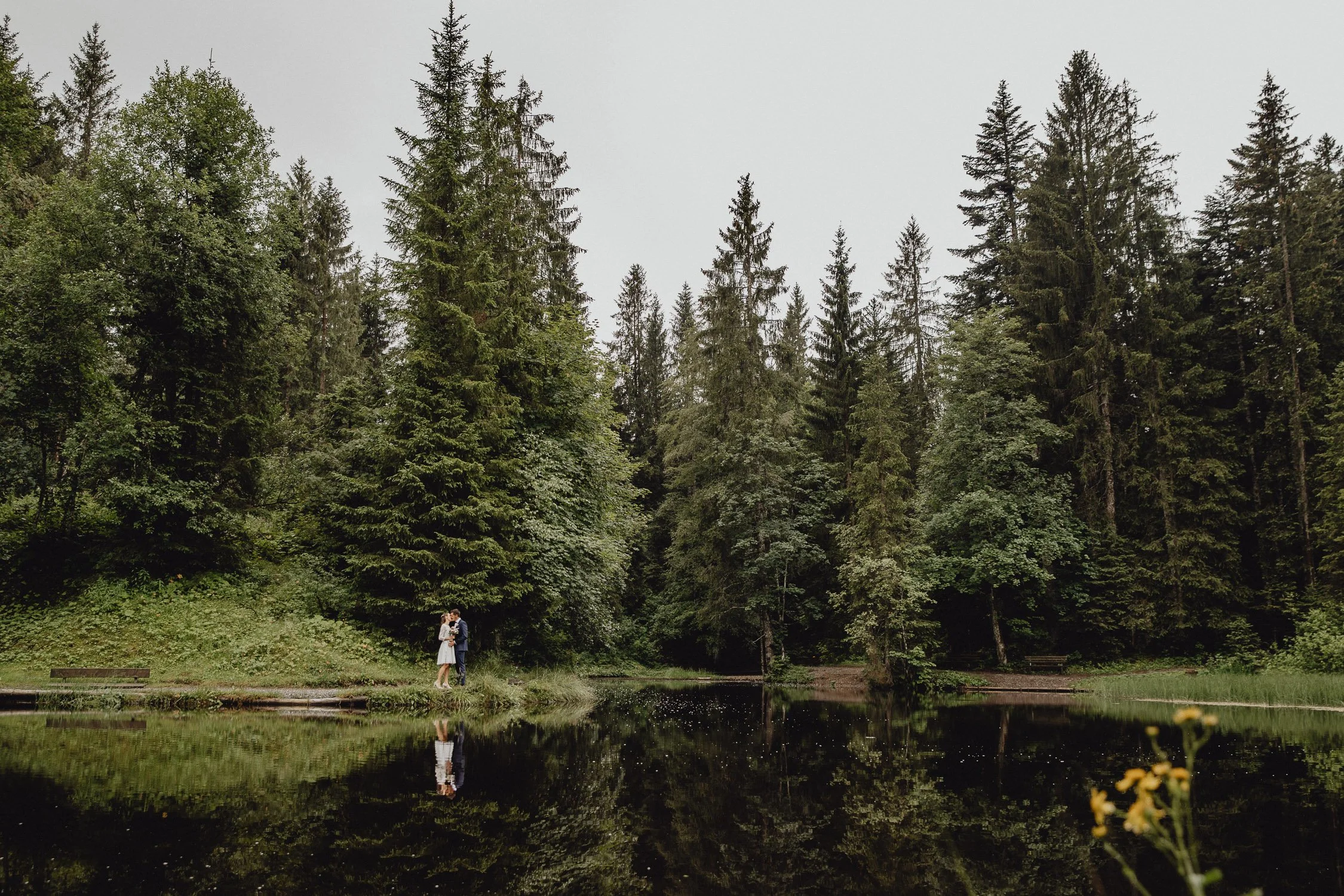 Ein Paar steht am Ufer eines Sees, umgeben von dichtem grünem Wald.