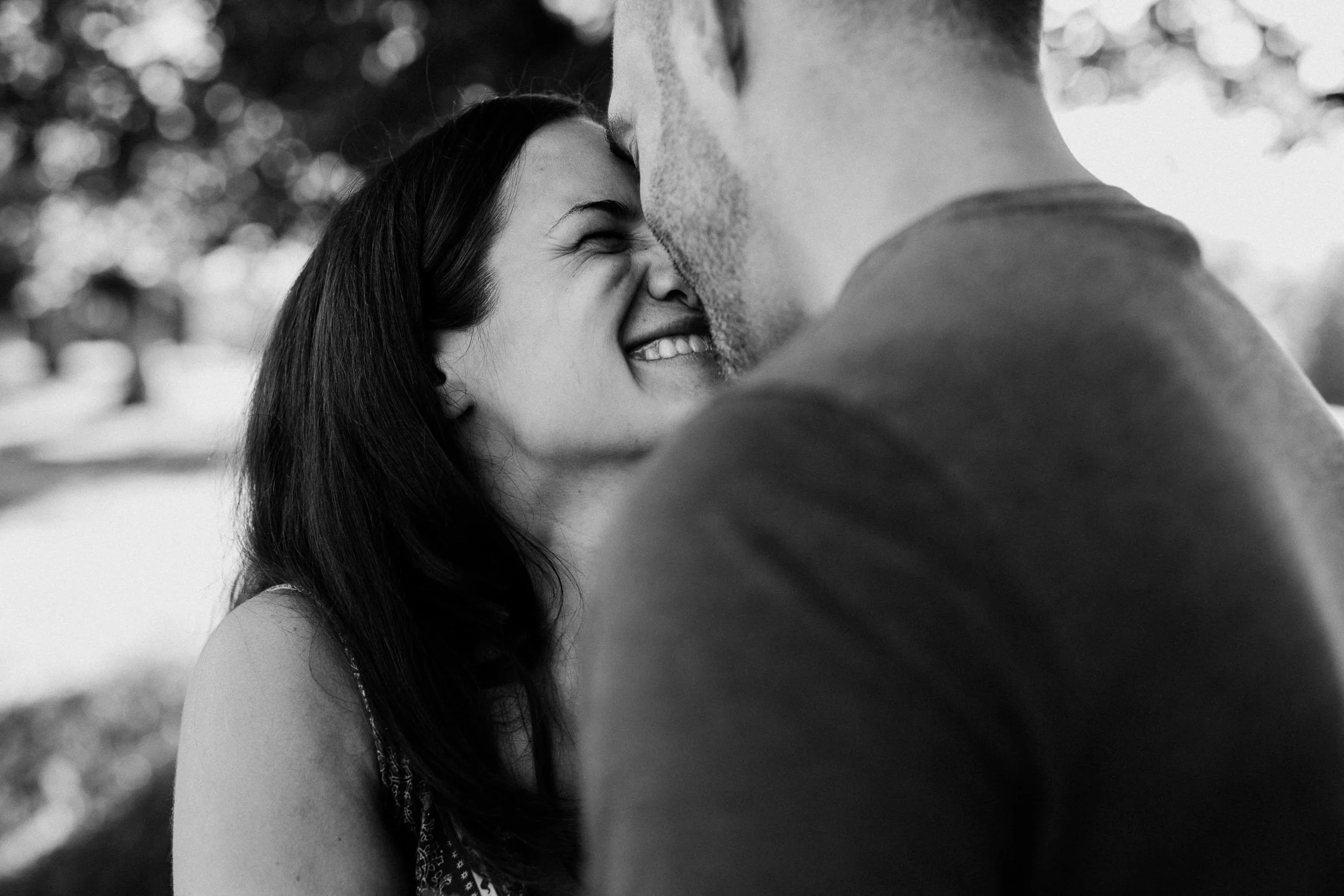 Ein glückliches Paar küsst sich zärtlich im Freien bei Sonnenlicht, umrundet von Bäumen, schwarz-weiß Fotografie.