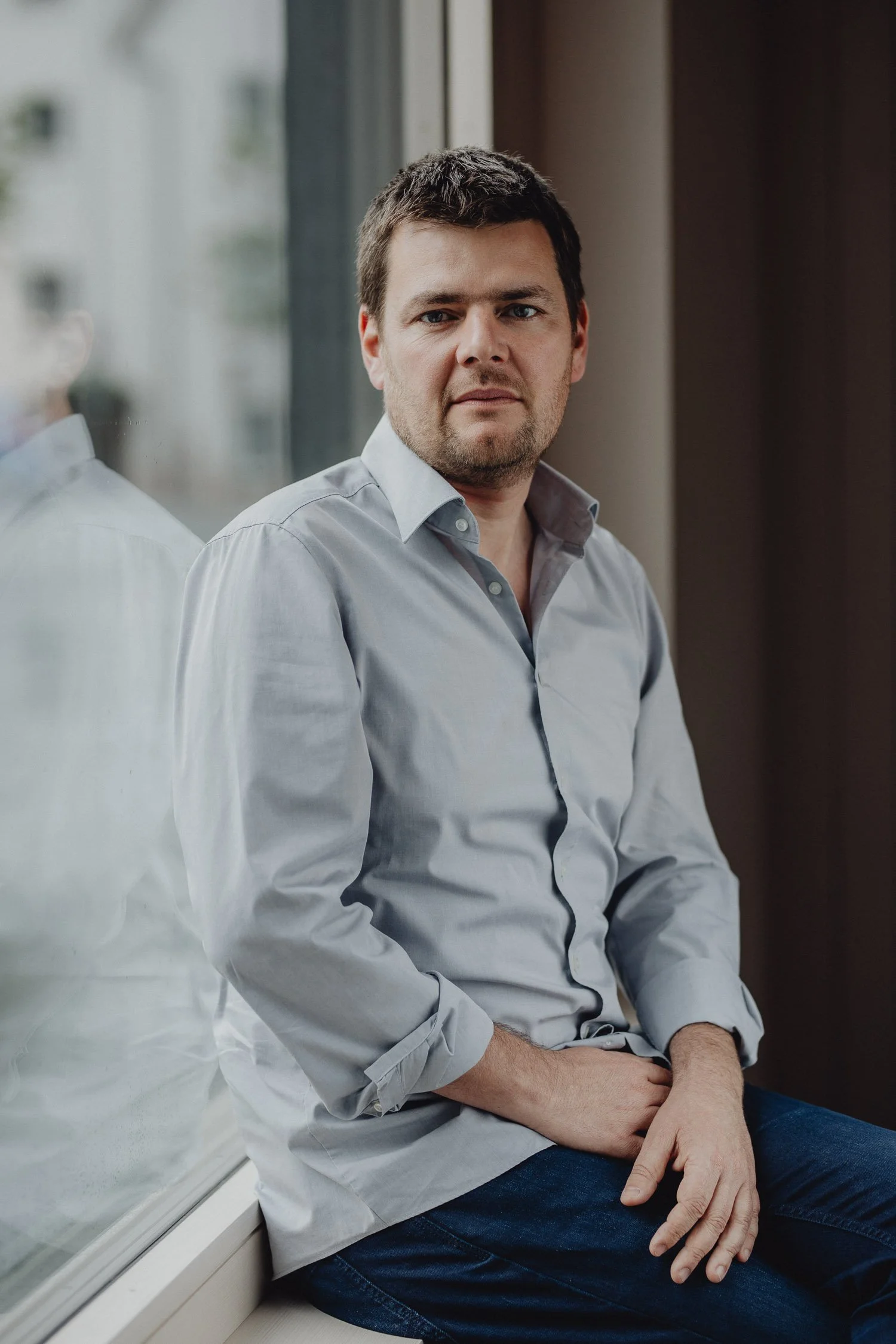 Ein junger Mann mit braunen Haaren und Bart sitzt an einem Fenster, trägt ein hellgraues Hemd und blaue Jeans, schaut nach vorne.