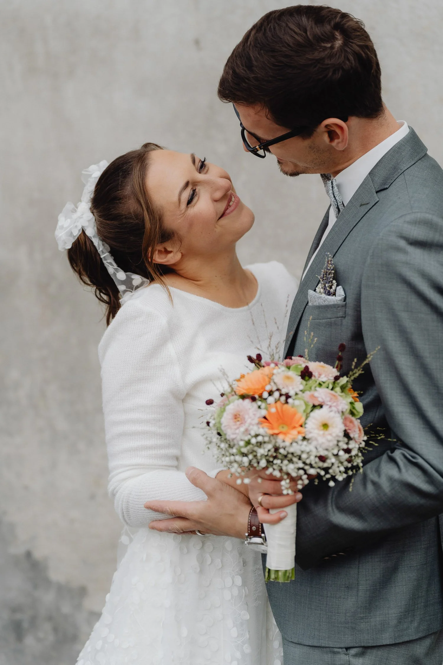 Ein Brautpaar, das sich auf einer Hochzeit liebevoll umarmt, mit der Braut, die einen bunten Blumenstrauß hält, vor einem neutralen Hintergrund.
