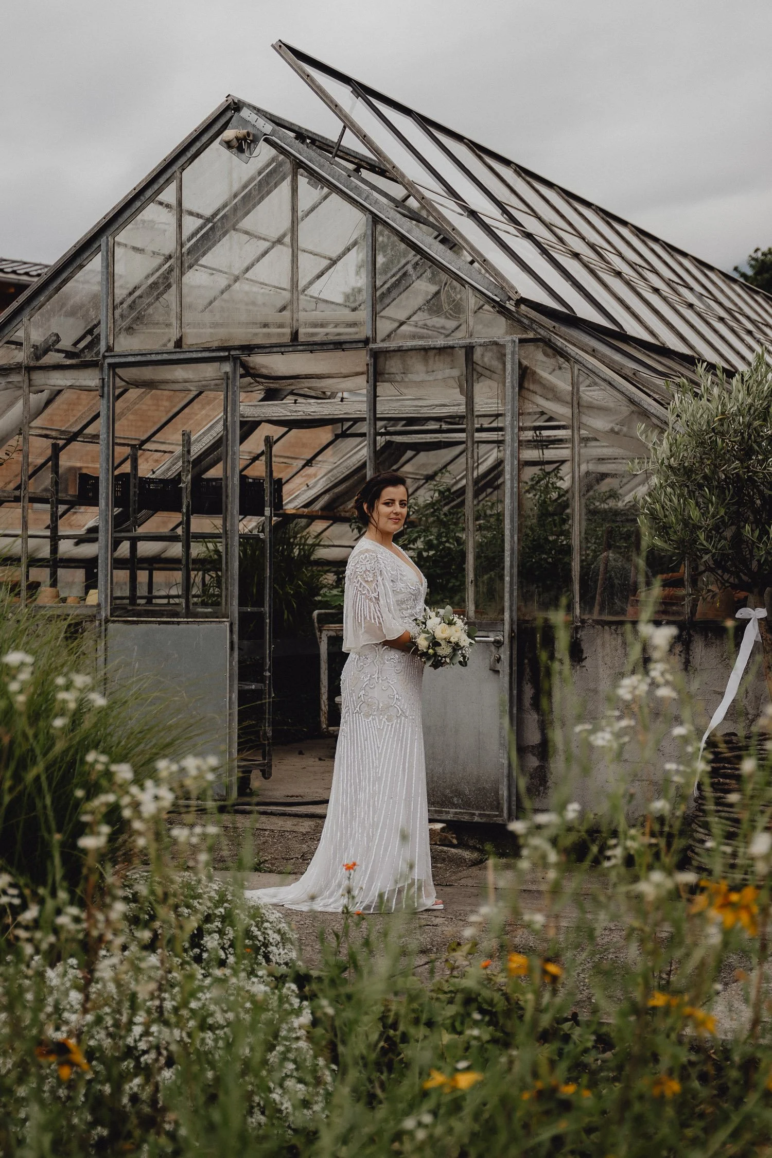 Eine Braut in einem weißen Hochzeitskleid steht vor einem alten Gewächshaus, hält einen Blumenstrauß und lächelt. Im Vordergrund sind Blumen und Gräser sichtbar, der Himmel ist bewölkt.