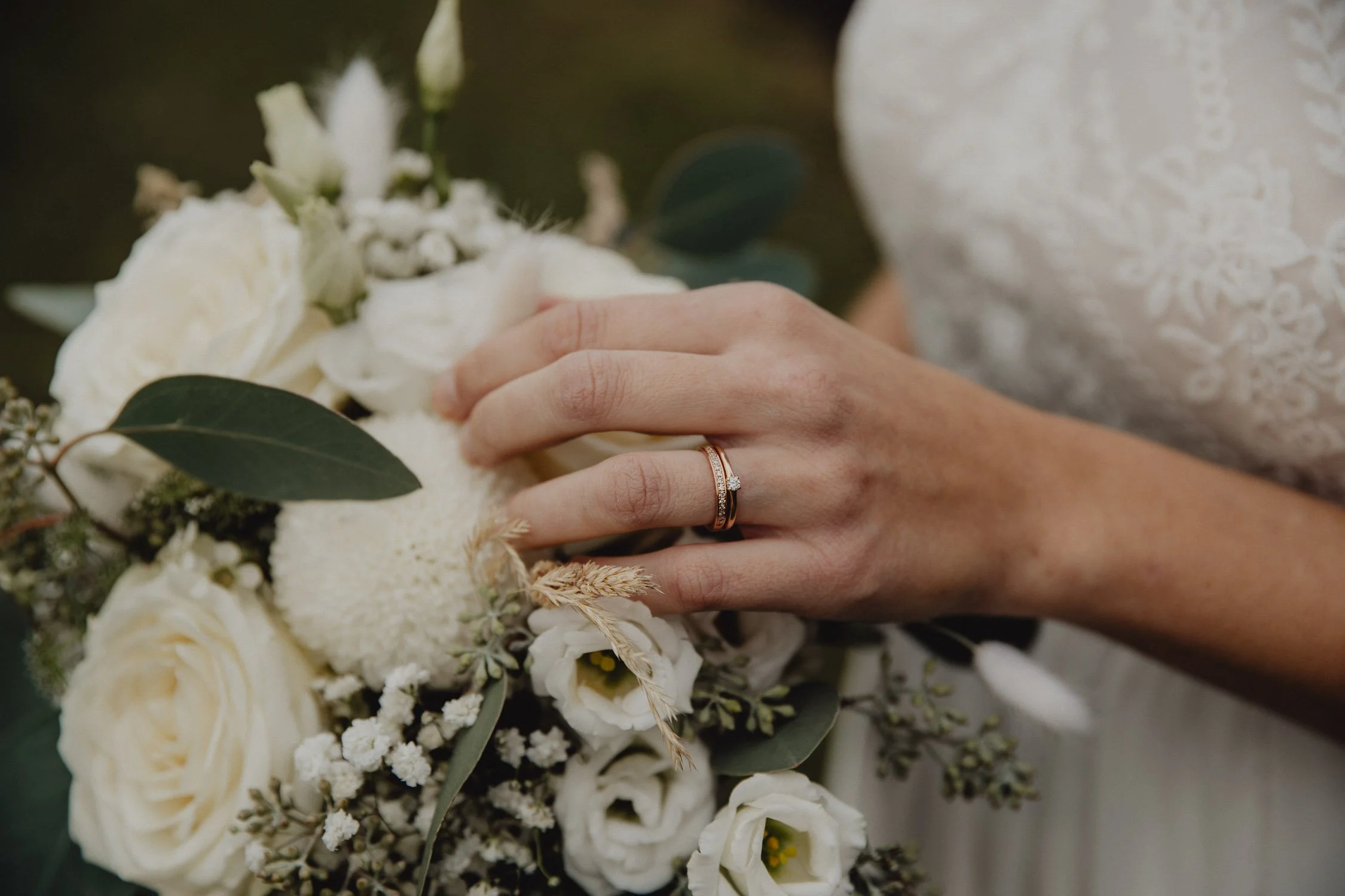 Nahaufnahme einer Hand mit Eheringen, die auf einem Brautstrauß liegt, bestehend aus weißen Rosen und anderen Blumen, während eine Braut in einem weiß-blumigen Kleid im Hintergrund sichtbar ist.