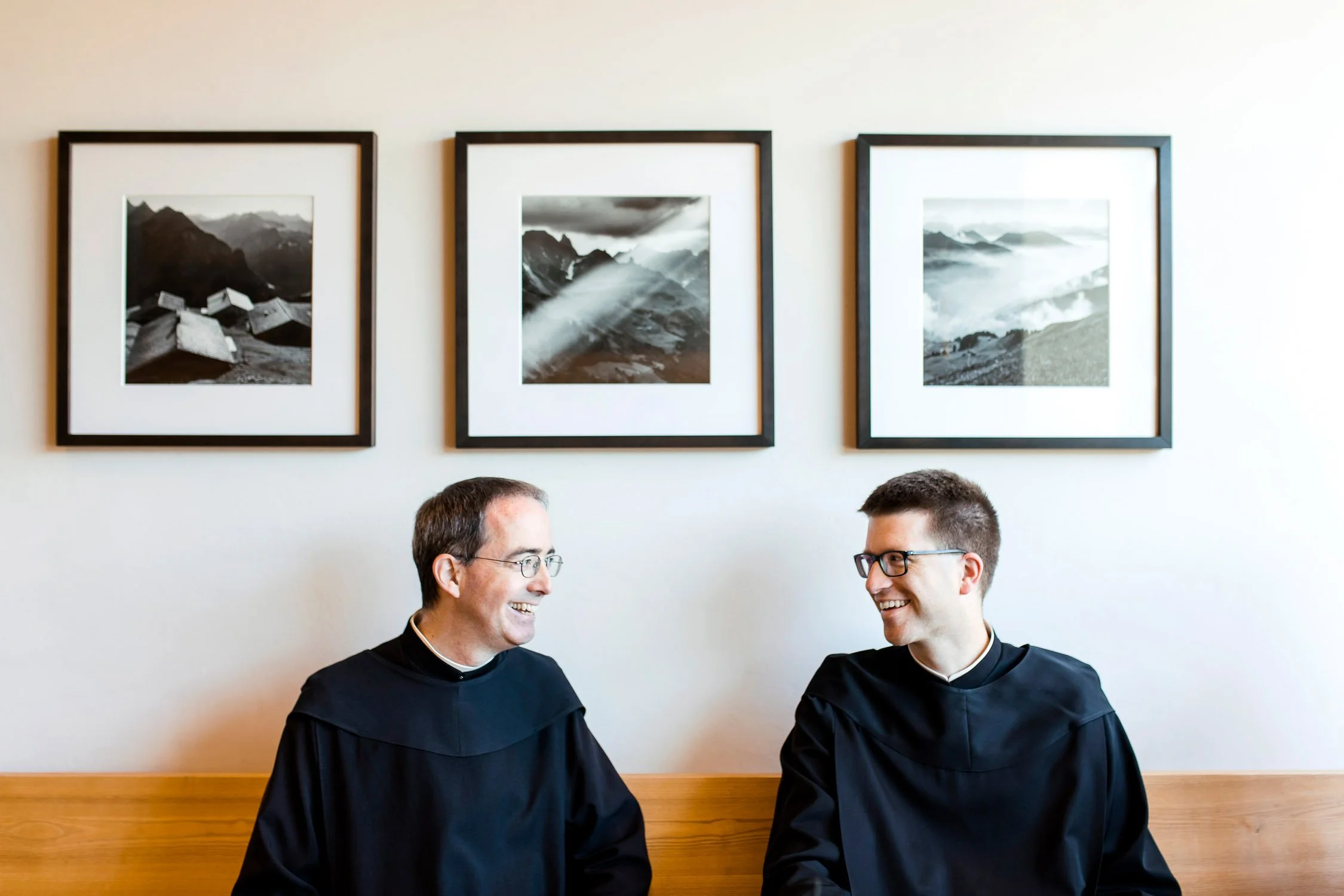 Zwei Männer in schwarzen Roben sitzen an einem Tisch und sprechen in einem Raum mit weißen Wänden und drei Schwarz-Weiß-Fotografien von Bergen im Rahmen.