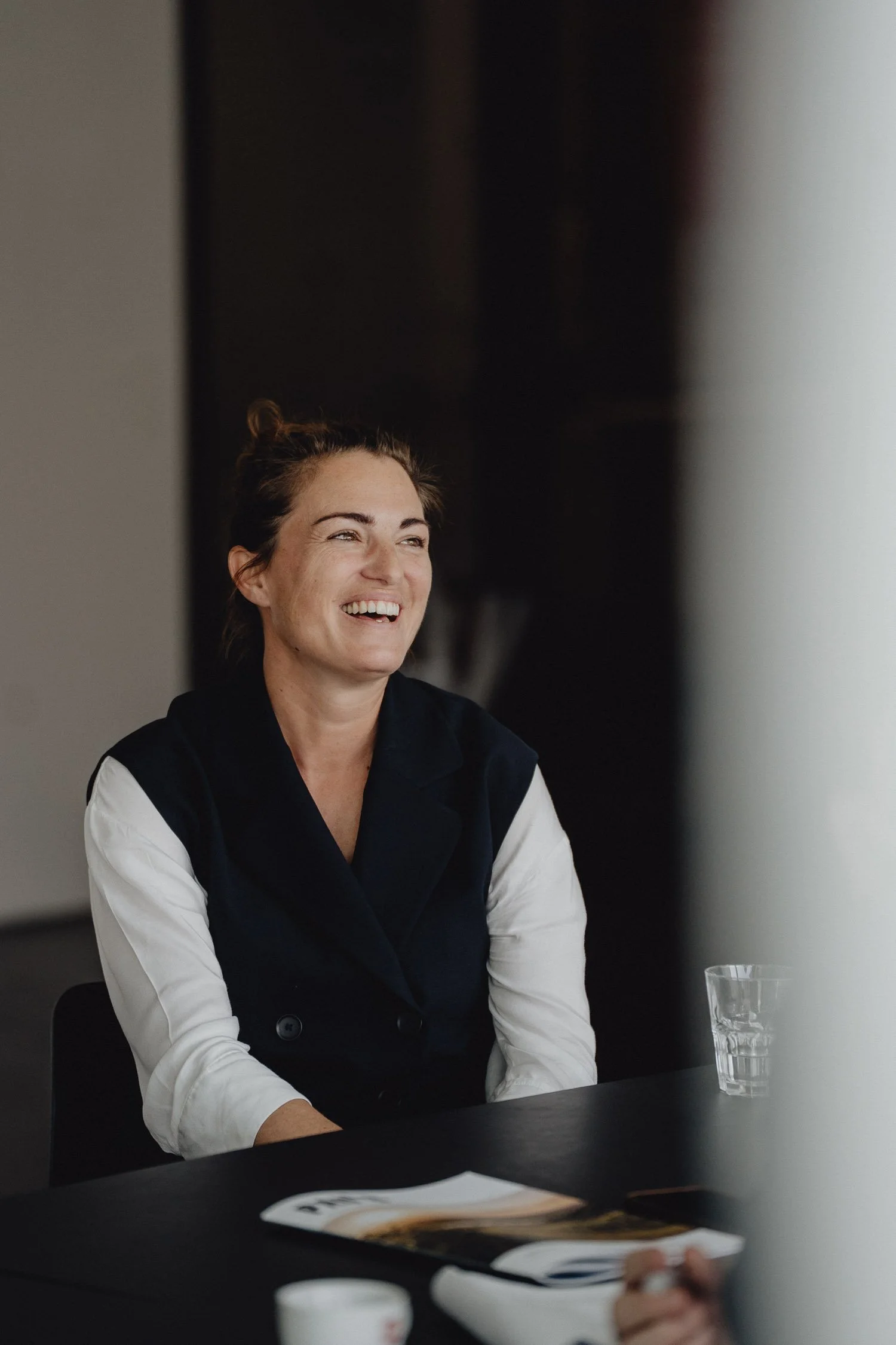 Eine lachende Frau mit braunen Haaren, die in einem Gespräch sitzt, bei einem Meeting oder Interview, mit einem Glas Wasser und Zeitschriften auf dem Tisch.