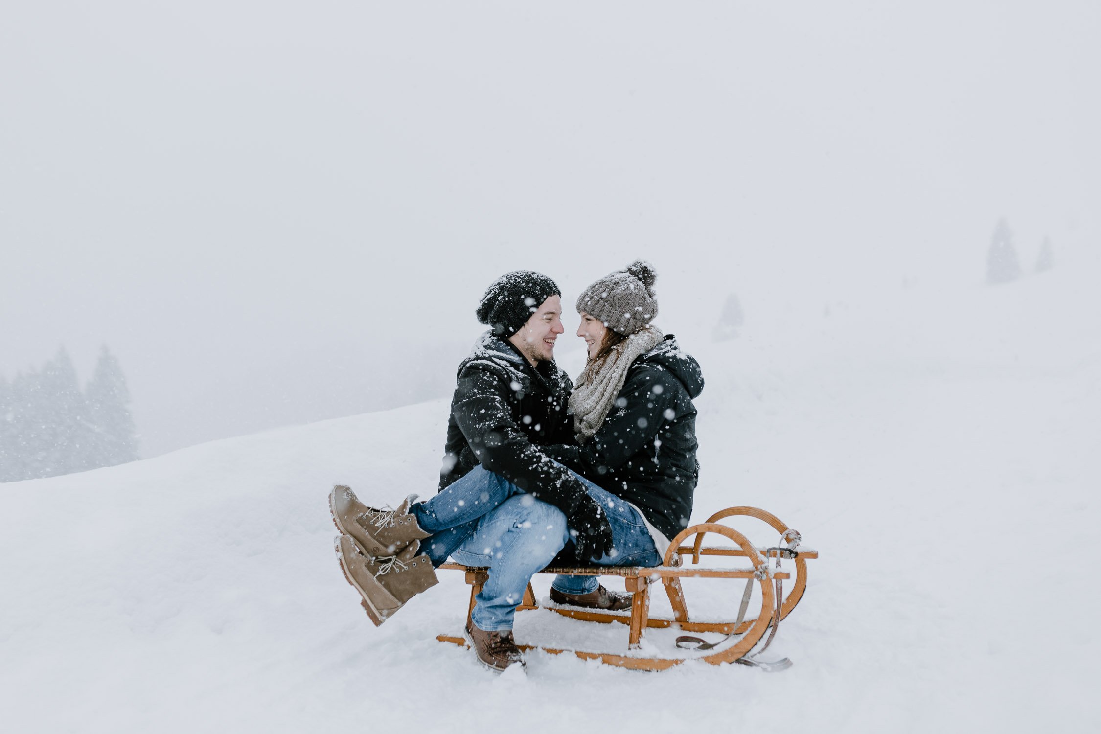 Ein junges Paar, das auf einem Schlitten im Schnee sitzt und sich liebevoll ansieht.