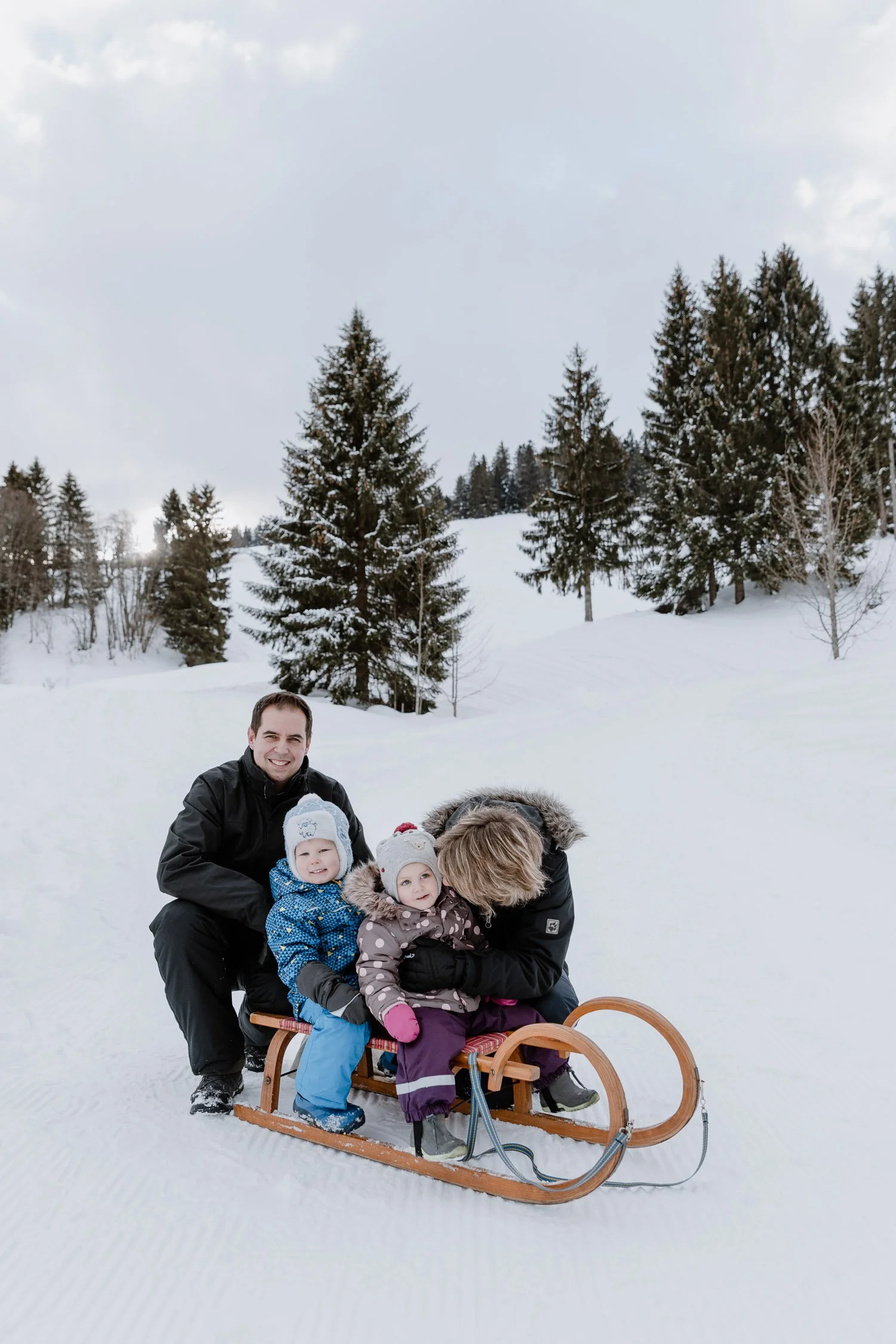 Familie mit zwei kleinen Kindern auf Rodel in verschneiter Winterlandschaft mit Tannenbäumen.