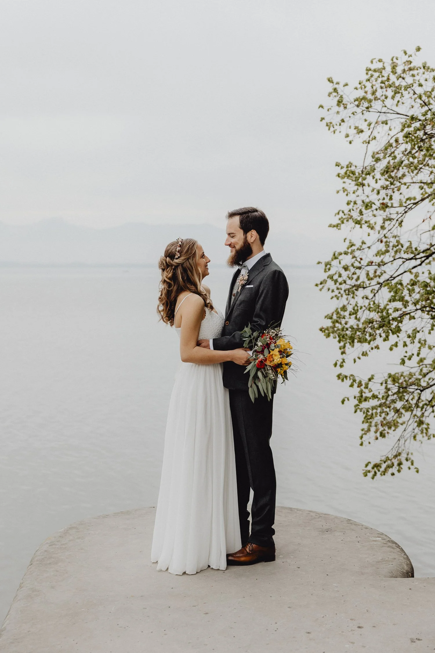 Ein Brautpaar steht auf einer Plattform am Wasser, blickt sich liebevoll an, die Braut trägt ein weißes Kleid mit Spaghetti-Trägern, der Bräutigam ein dunkles Anzug, das Mädchen hält einen bunten Blumenstrauß.