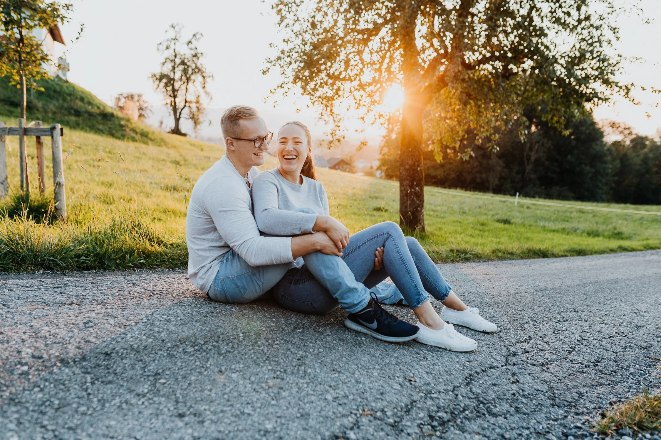 Ein lachendes Paar sitzt auf einer Straße im Freien bei Sonnenuntergang, umgeben von Bäumen und grünem Gras.