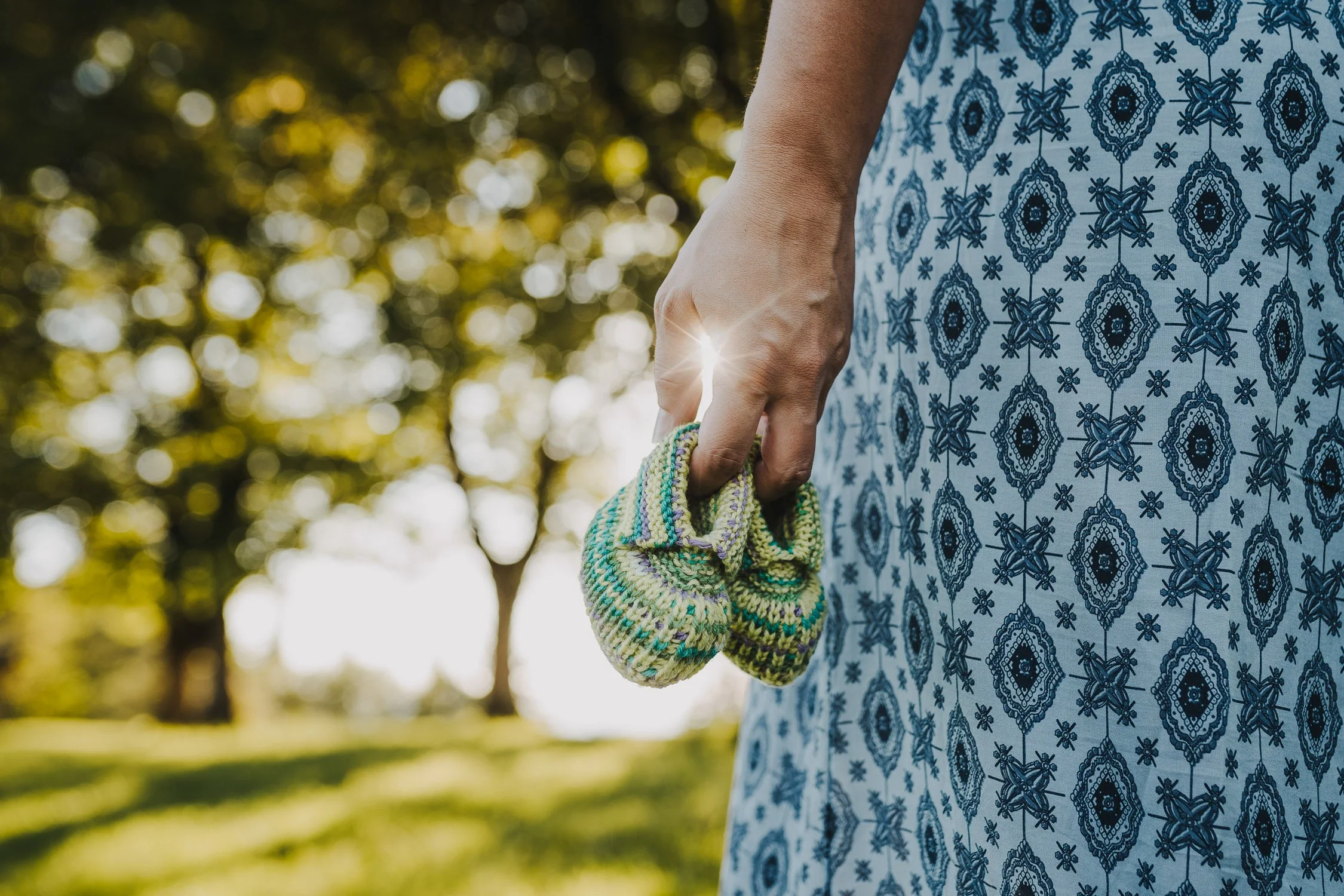 Nahaufnahme einer Frau, die kleine bunte Babyschuhe in der Hand hält, im Hintergrund Bäume und Sonnenlicht.